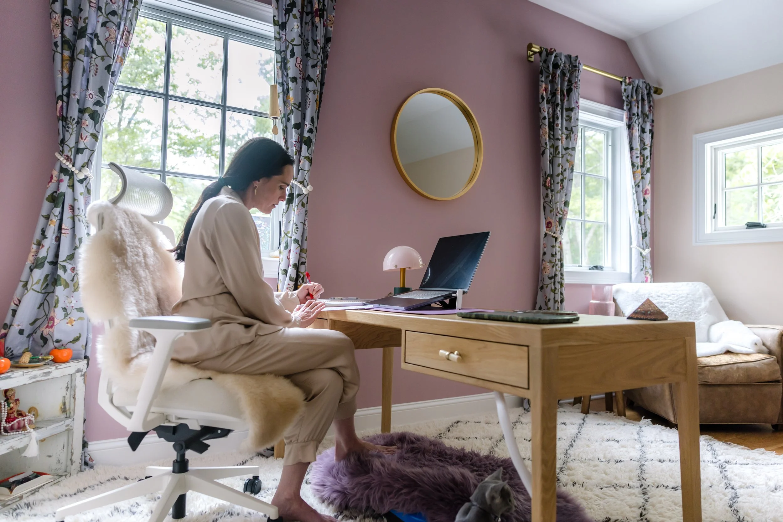 A woman sitting in a white office chair at a wooden desk, working on a laptop, in a room with pink and cream walls, floral curtains, and a mirror on the wall. There is a small table with snacks and a cat lying on a fluffy purple rug under her feet.