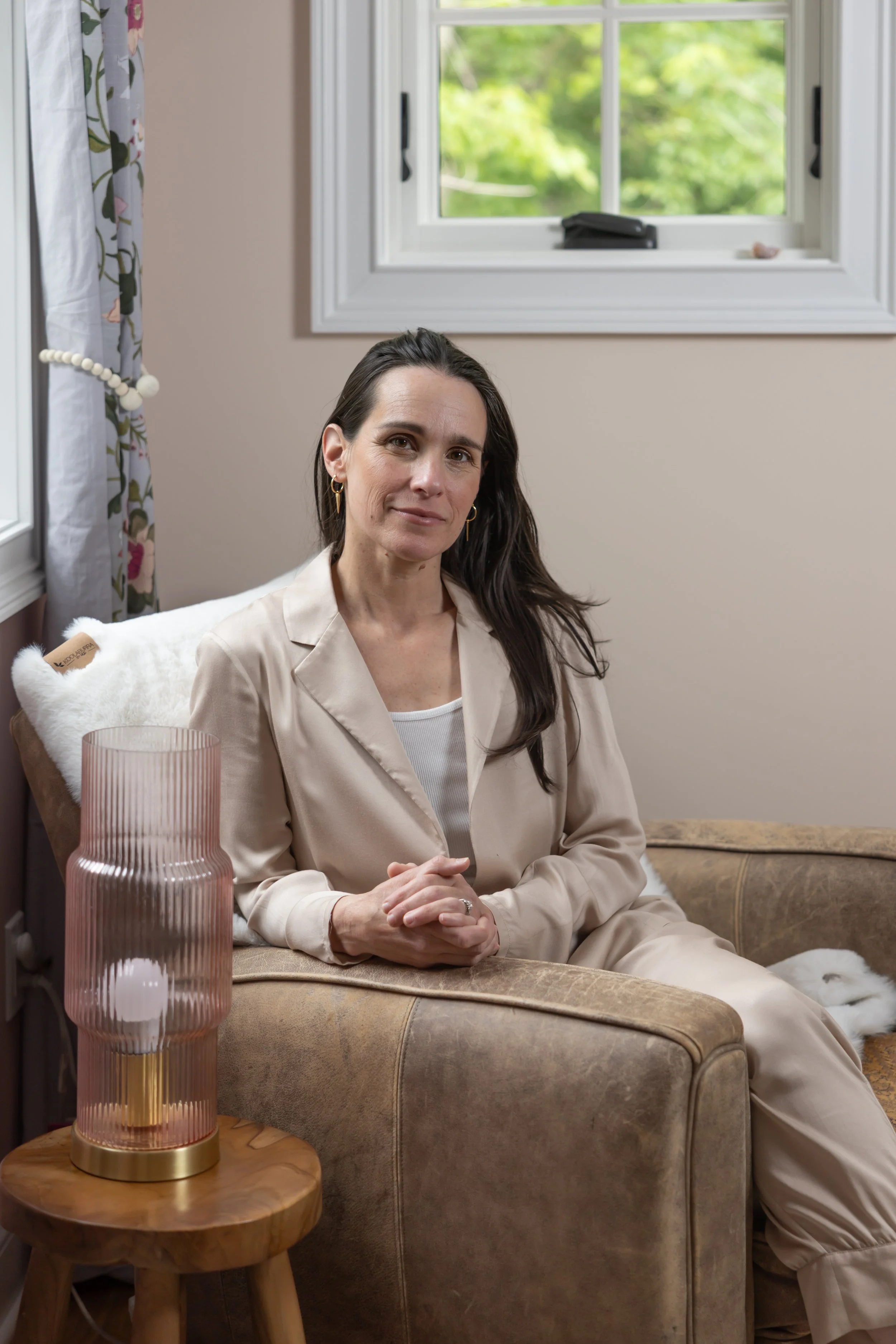 A woman with long dark hair, gold hoop earrings, and beige blazer sitting on a brown couch in a cozy room with a window behind her showing green trees outside. There is a pink ribbed lamp on a wooden side table next to her.