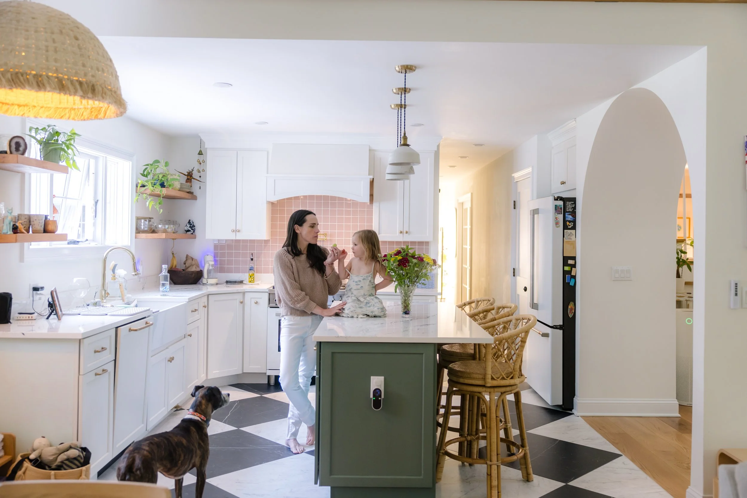 A woman and a young girl standing in a bright kitchen, with the woman giving a snack to the girl. The kitchen has white cabinets, pink backsplash, and a large island with green base and wooden chairs. There are plants on the windowsill and shelves, a dog in the foreground, and sunlight streaming through the windows.