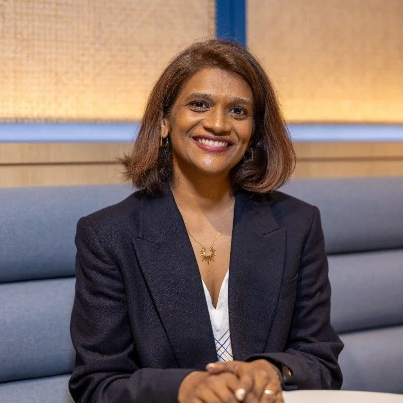 Smiling woman with shoulder-length brown hair, wearing a dark blazer and a gold sunburst necklace, sitting on a blue couch in a professional setting.