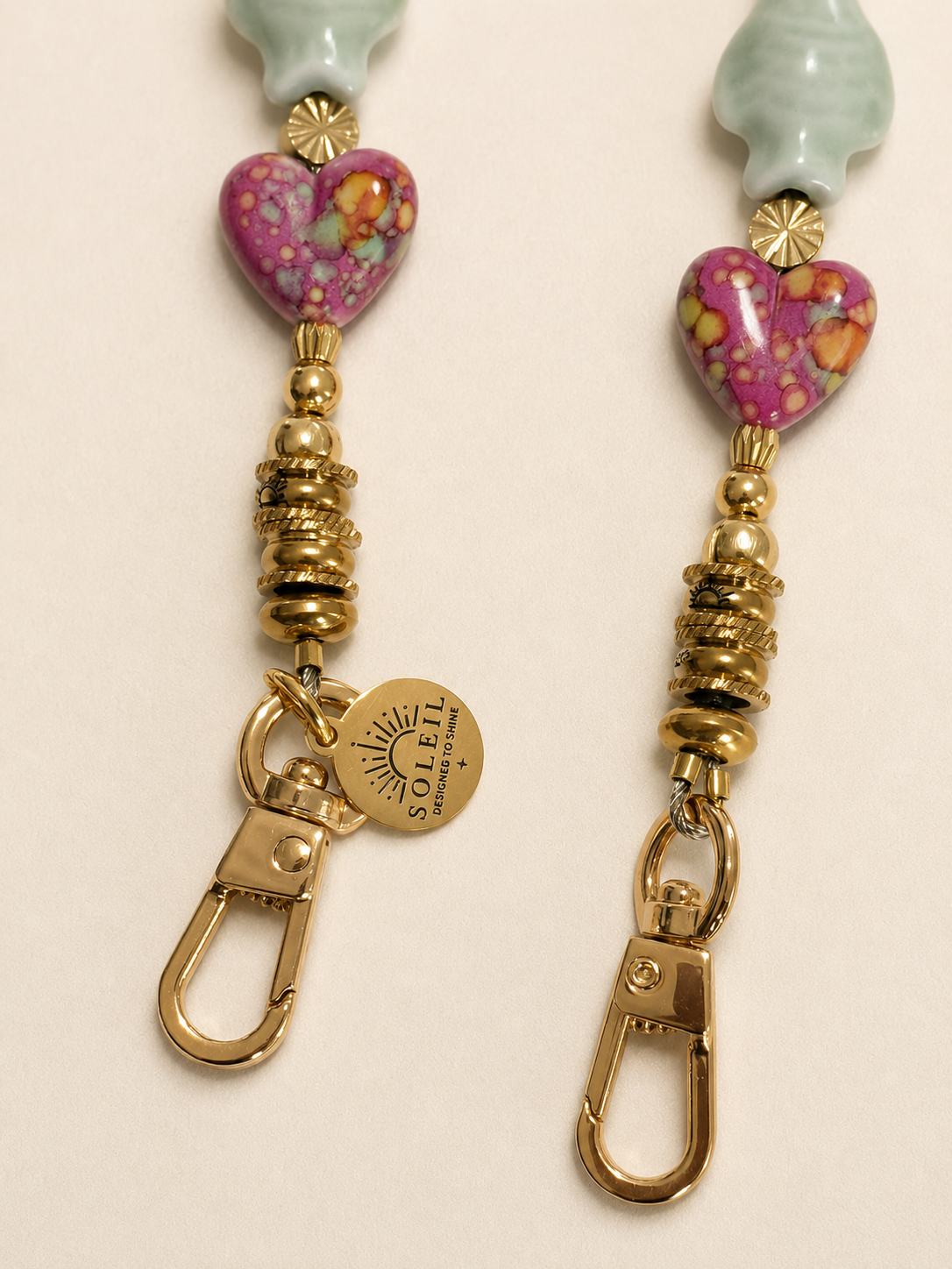 Close-up of two gold keychains with beaded and heart-shaped glass pendants, featuring pink, yellow, and green patterns, on a light background.