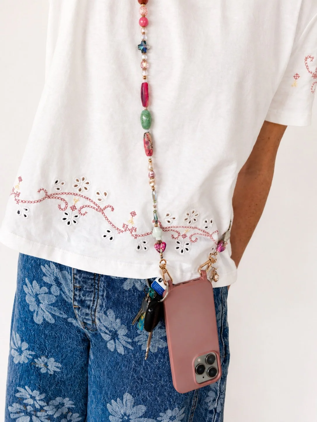 Person wearing a white embroidered top with pink and red beads, blue floral jeans, with a pink phone case attached to a lanyard around their neck, holding keys and key fob.