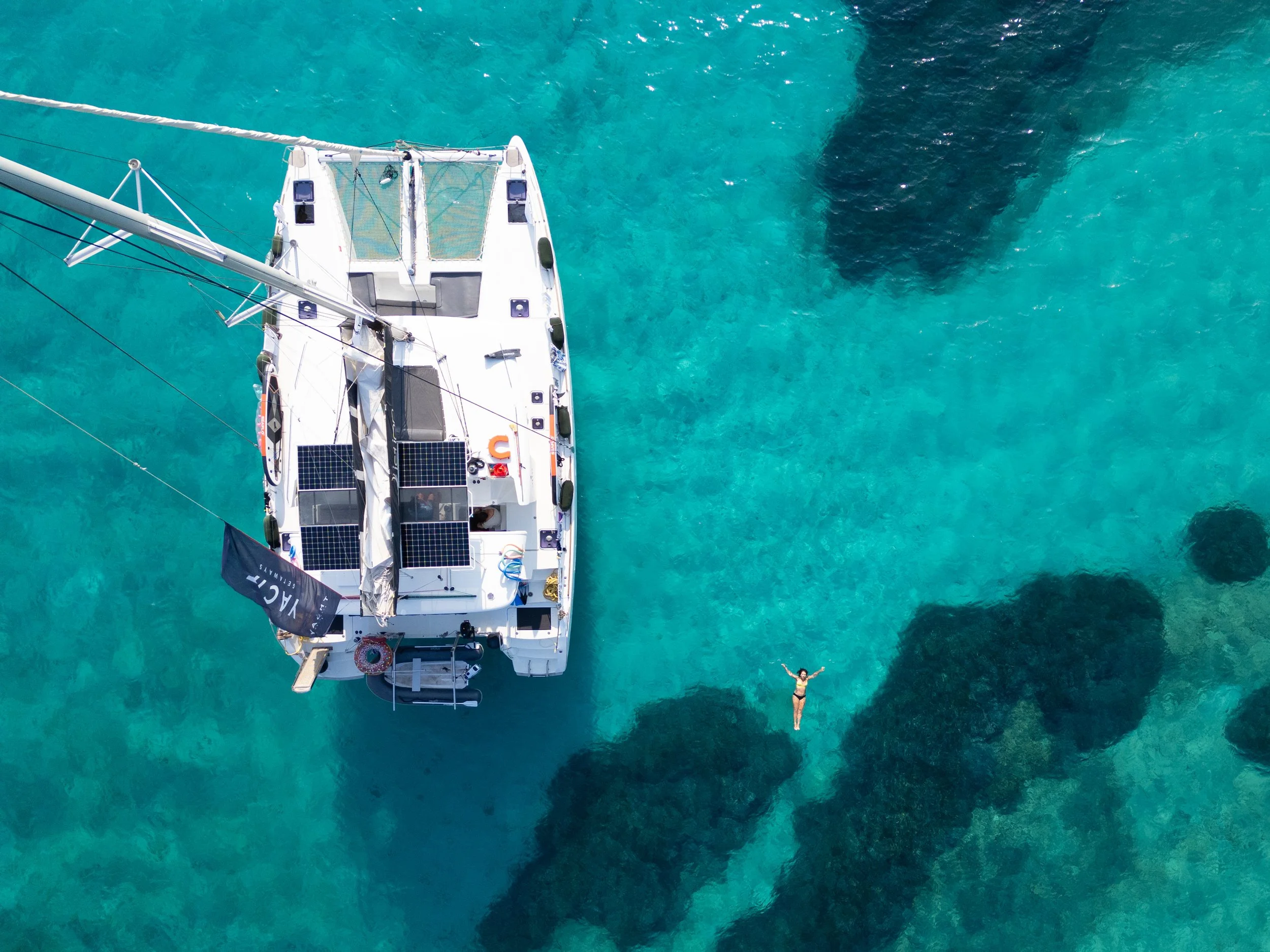 An aerial view of a white sailboat anchored in clear blue water with a woman swimming nearby, wearing a black bikini and sunglasses, enjoying a sunny day.