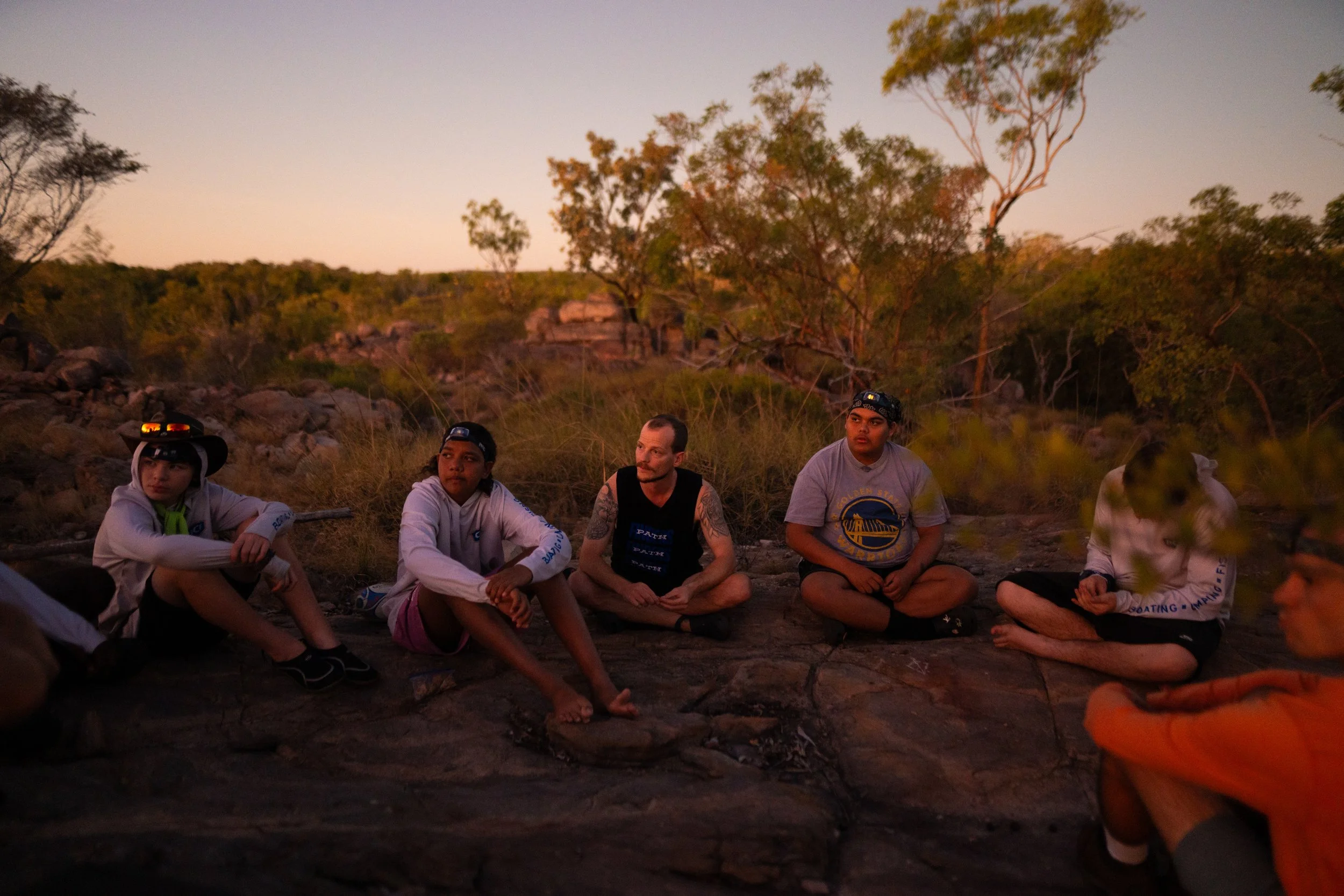 Group of five young people sitting on the ground outdoors during sunset, surrounded by trees and rocks.