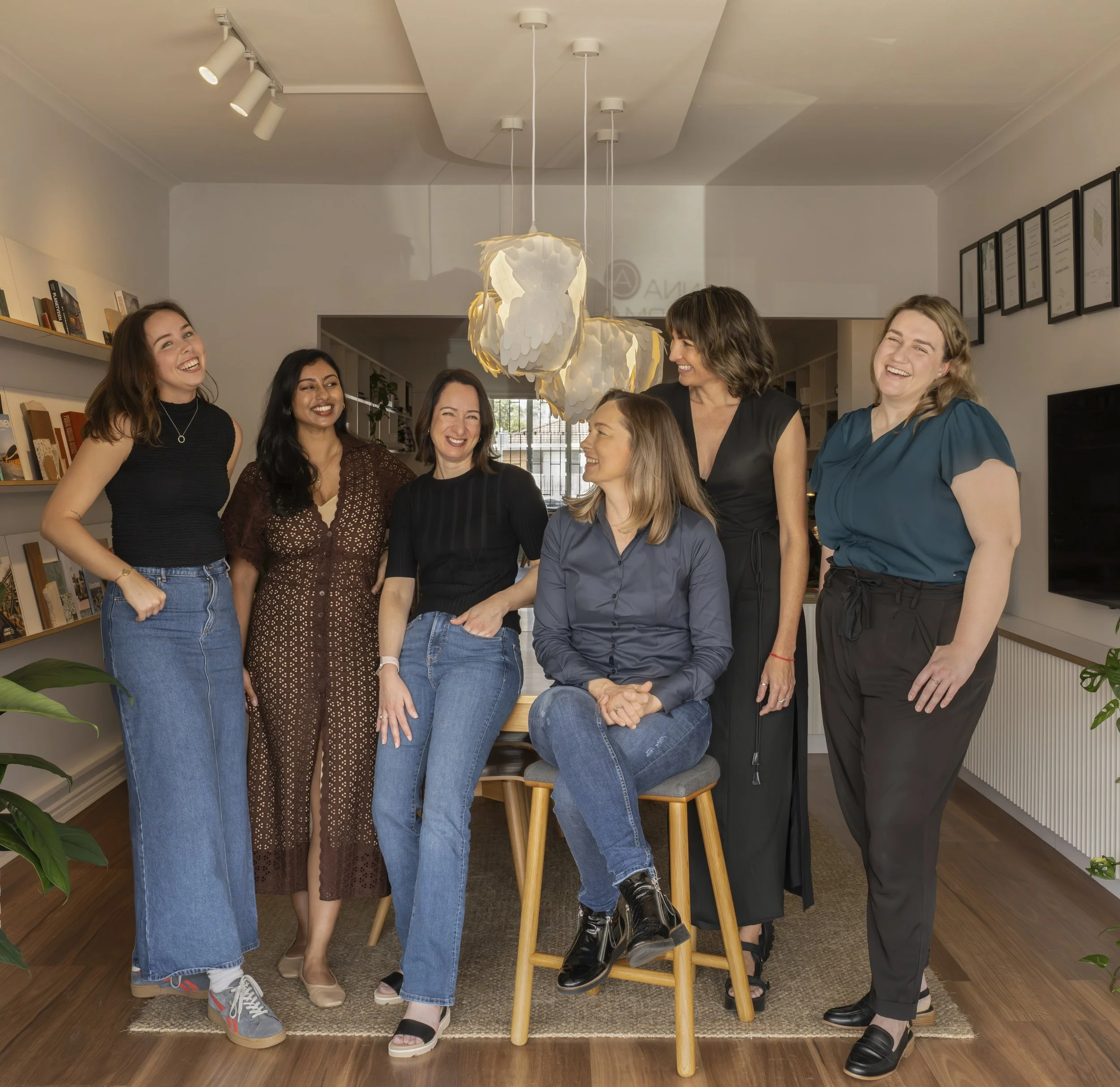 A diverse group of women gathered indoors, smiling and laughing, standing around a wooden stool, with a modern chandelier hanging from the ceiling, and framed documents on the wall.