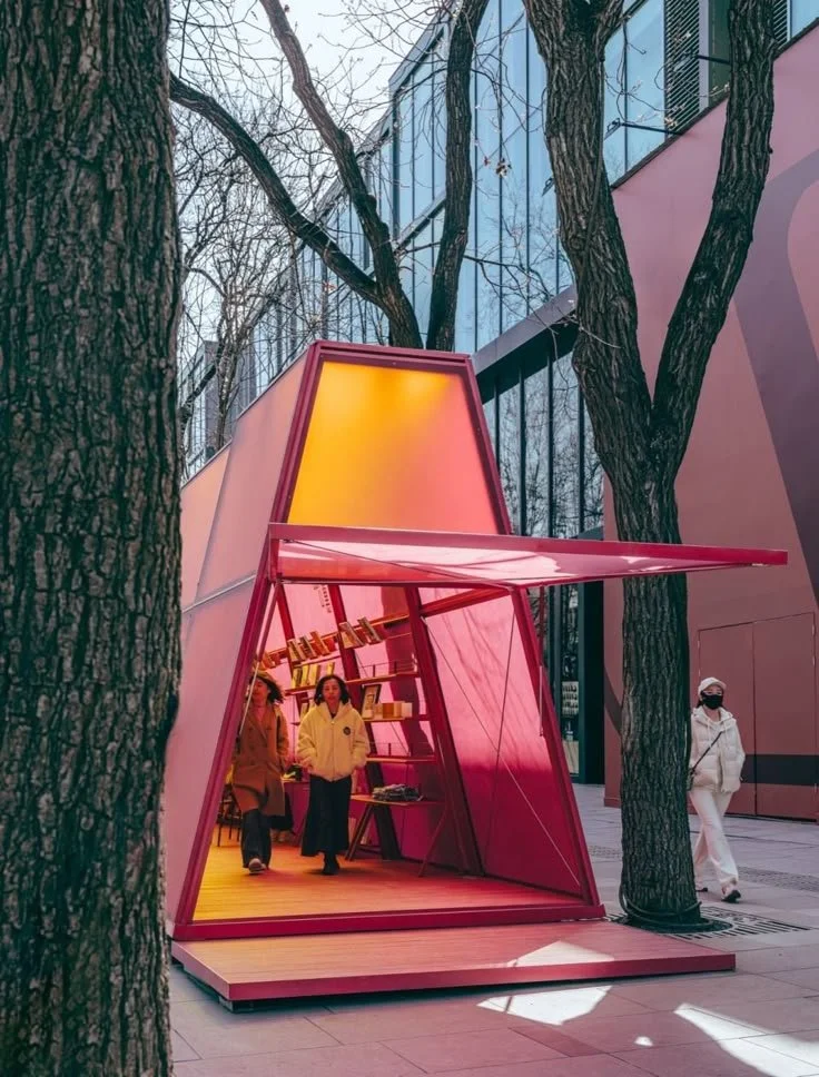 People inside a pink, angular, open-fronted structure with shelves, set against a modern building and trees on a city sidewalk.