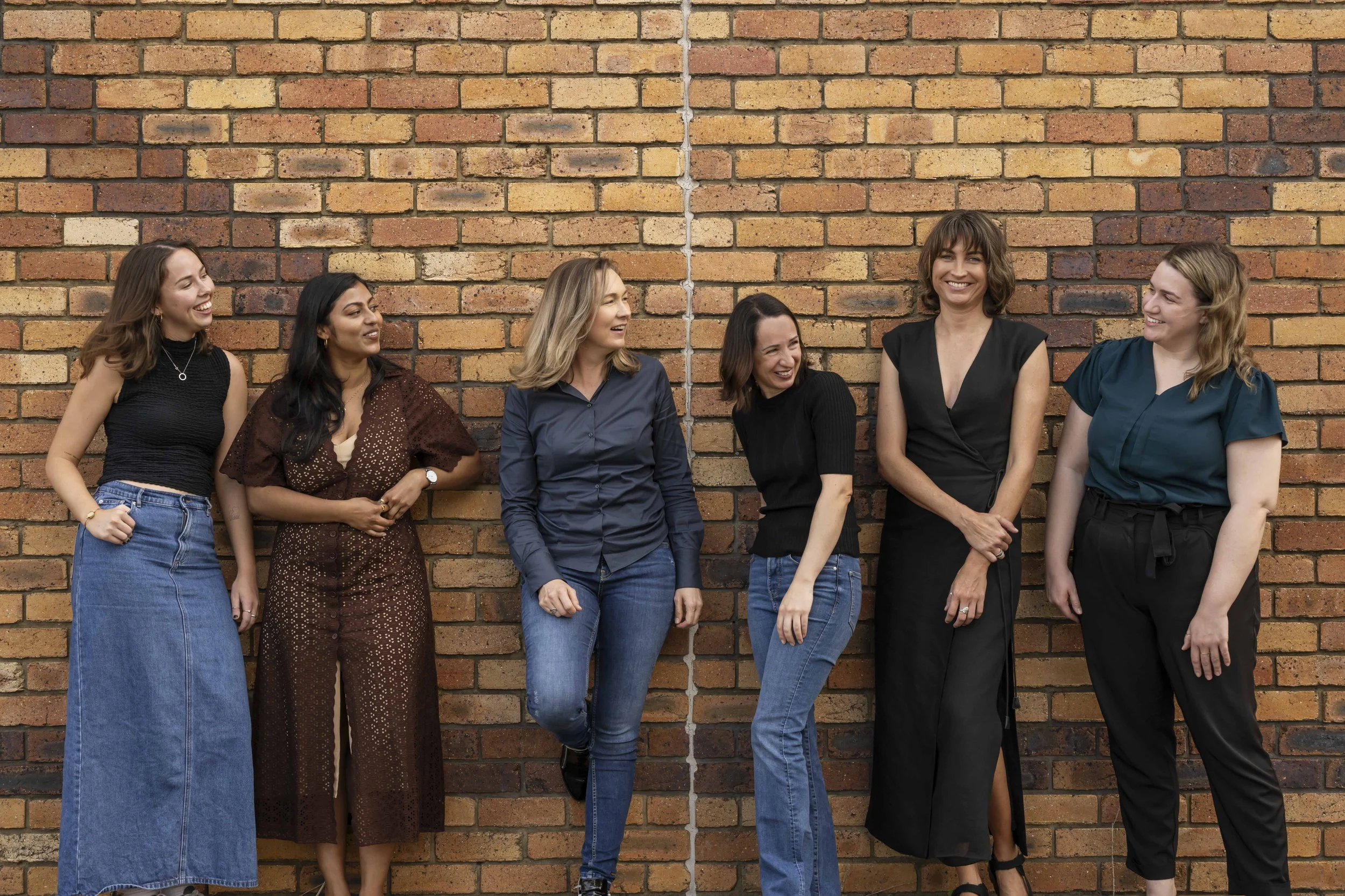 Six women standing and laughing against a brick wall, engaging with each other.