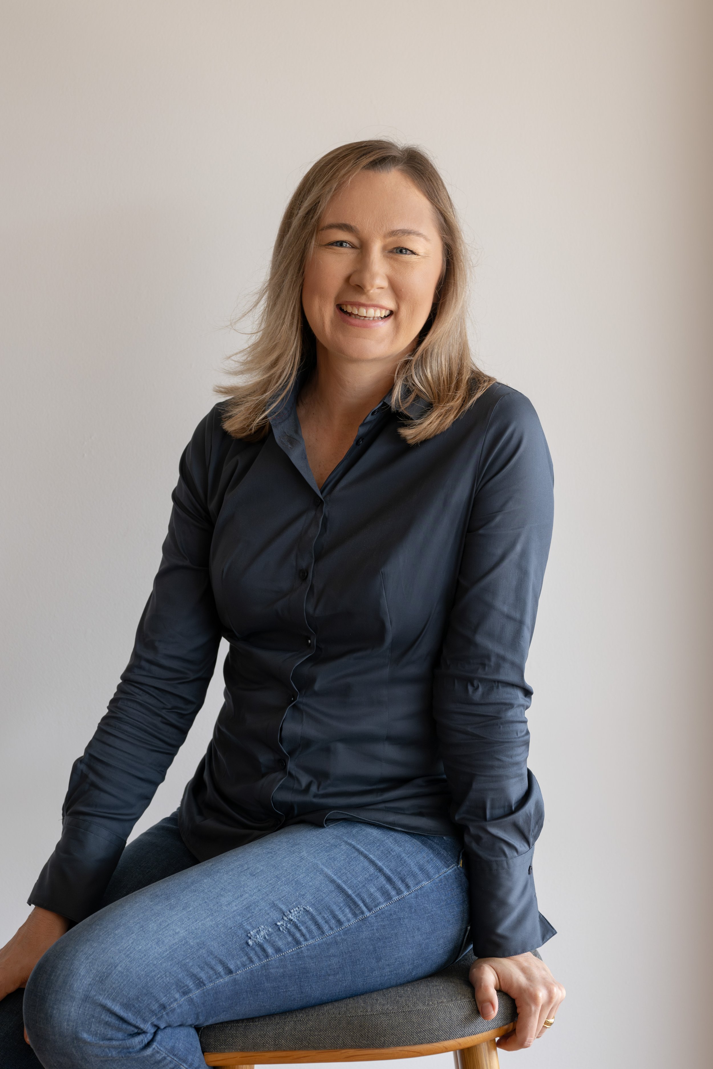 A woman with blonde hair, wearing a black satin shirt, sits on a stool with a smile. She is leaning slightly forward, with her right hand on her lap and left hand resting on the stool, against a plain light-colored wall.