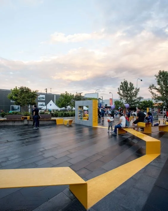 People sitting and walking in an outdoor urban plaza with yellow benches, trees, and a sky with clouds at sunset.