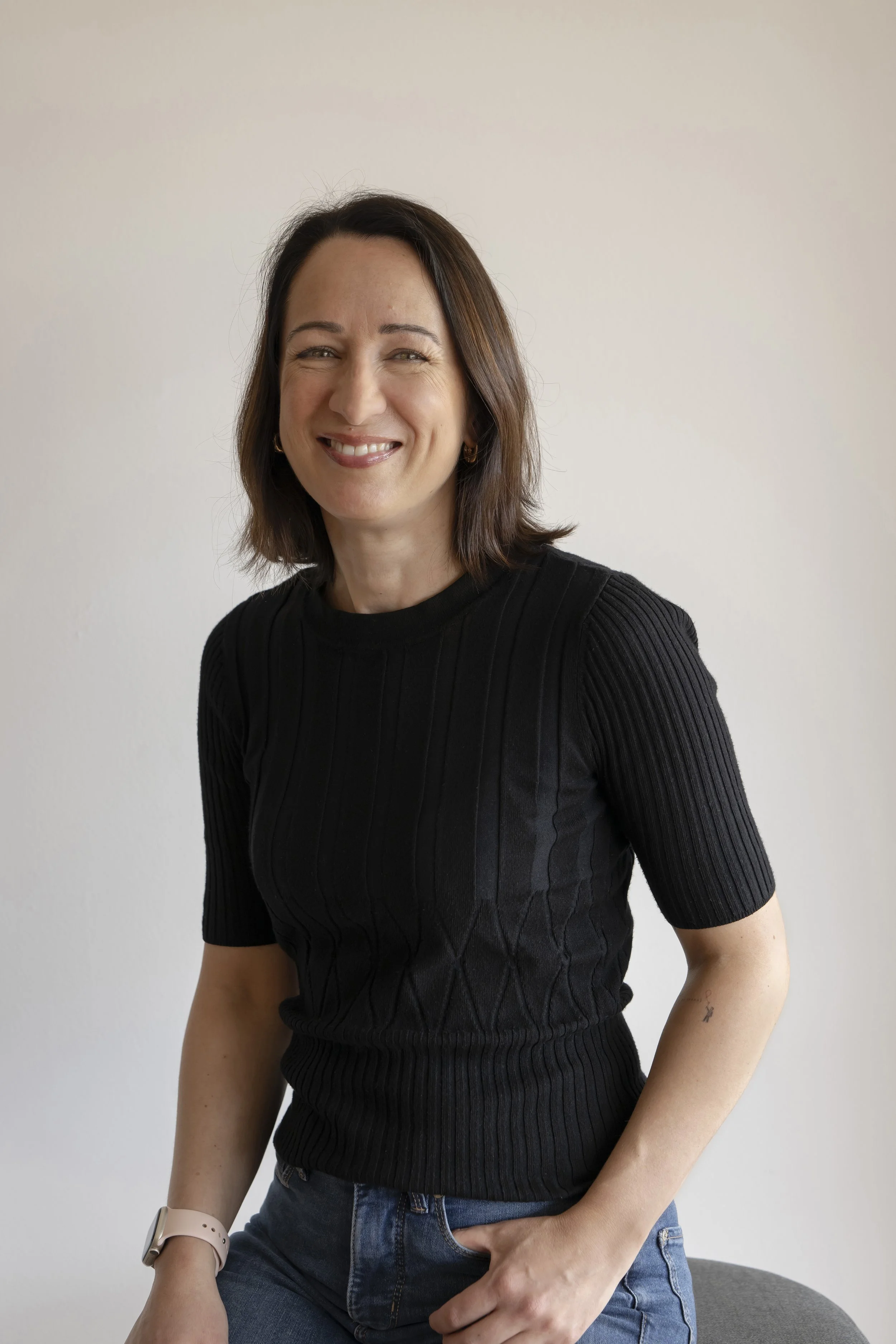 A woman sitting on a gray chair, wearing a black ribbed shirt, blue jeans, a smartwatch, and a light-colored wristband, smiling with her teeth showing and her hand resting on her lap. The background is plain white.