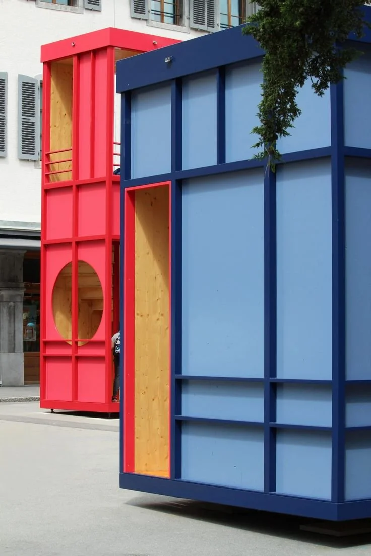Colorful outdoor art installation with red and blue structures, featuring geometric shapes and rectangular frames, in an urban setting.