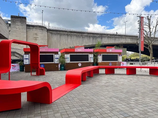 Outdoor seating area with red curved benches and ticket booths under a bridge at the National Theatre, with string lights overhead and a tree on the right.
