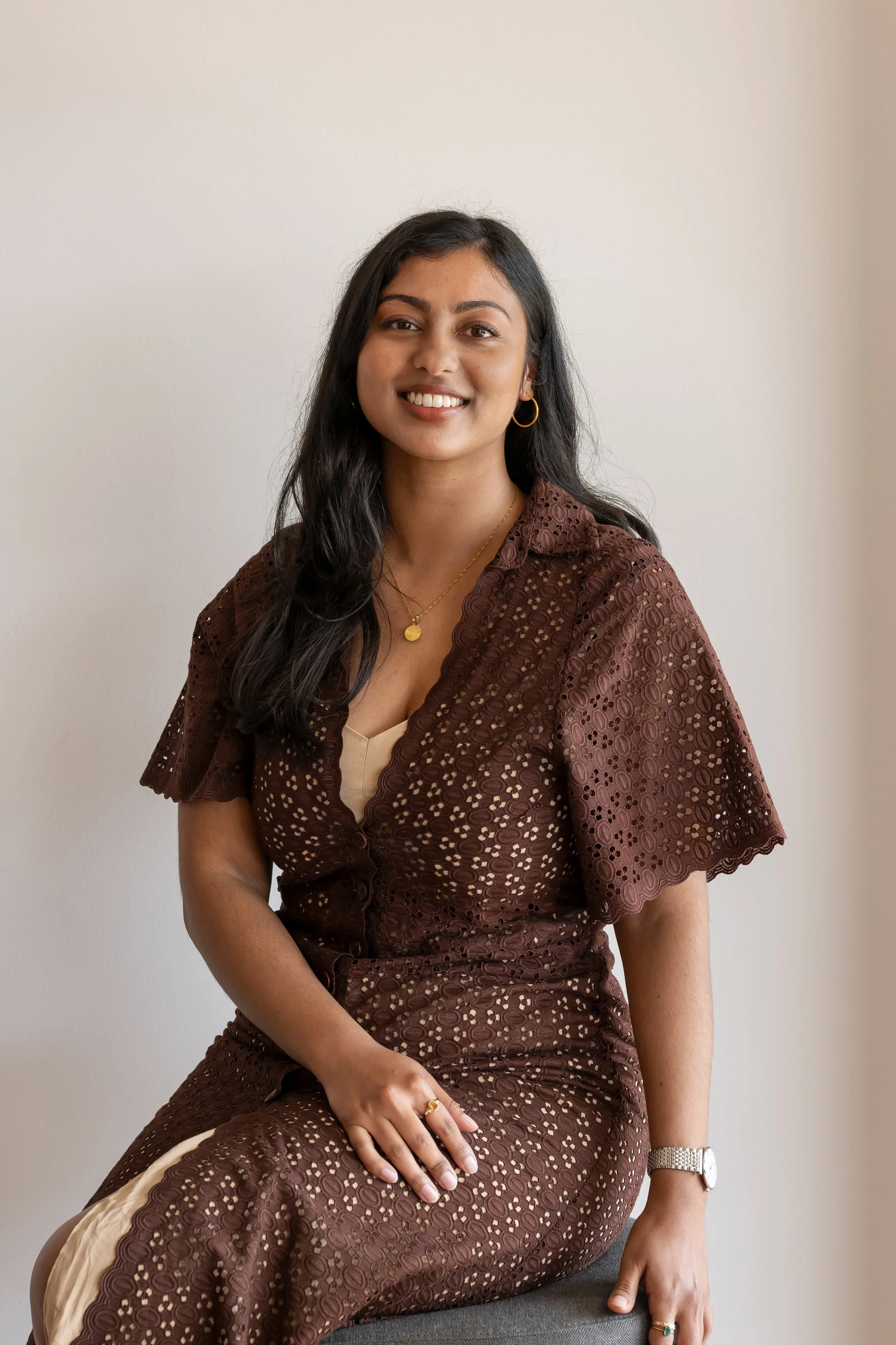 A smiling woman with long black hair wearing a brown lace dress with a beige underlay, sitting on a gray chair against a plain off-white wall. She accessorizes with gold jewelry and a silver watch.