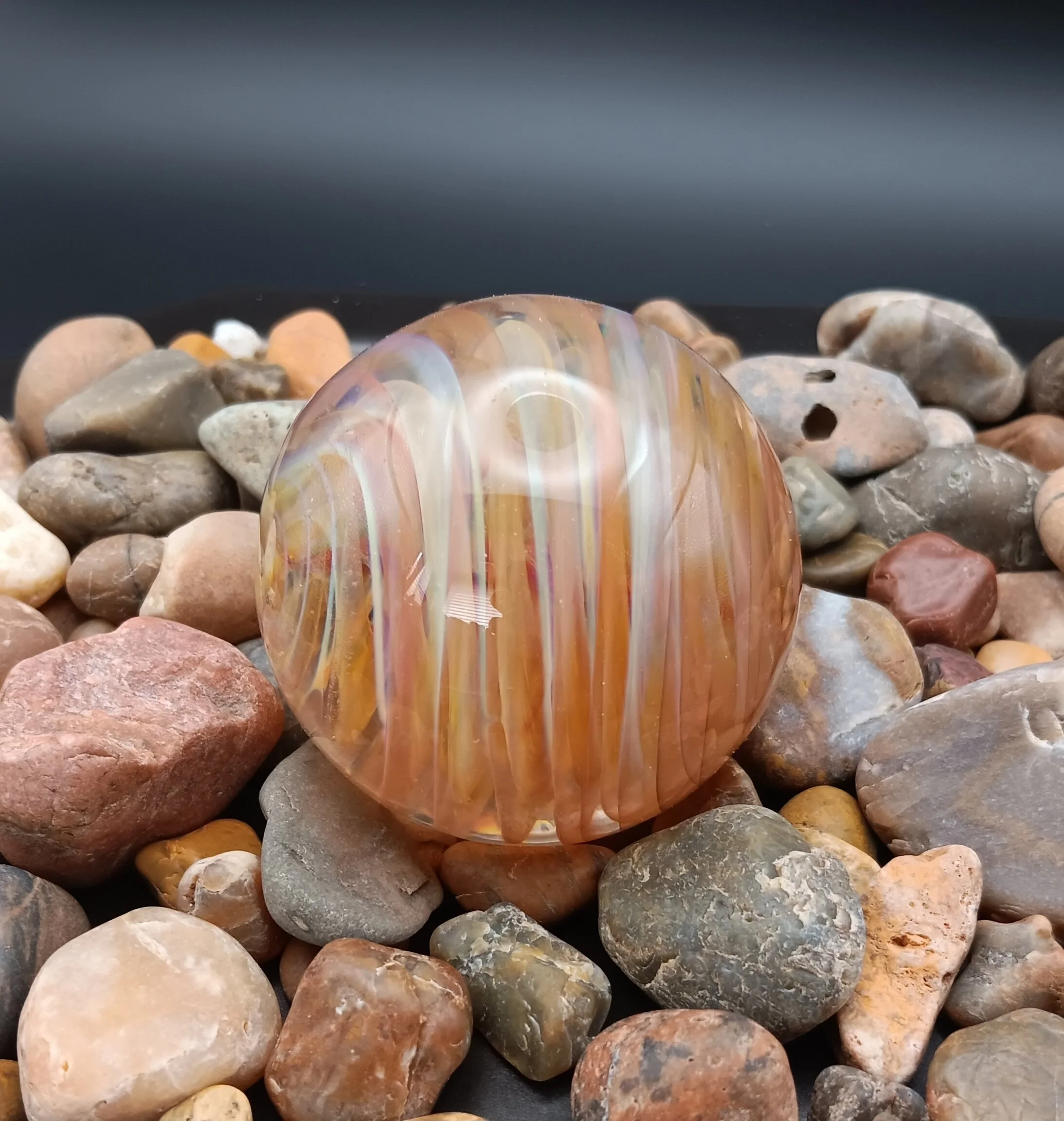 Close-up of multicolored, smooth, rounded stones and one orange glass marble with rainbow reflections among the stones.