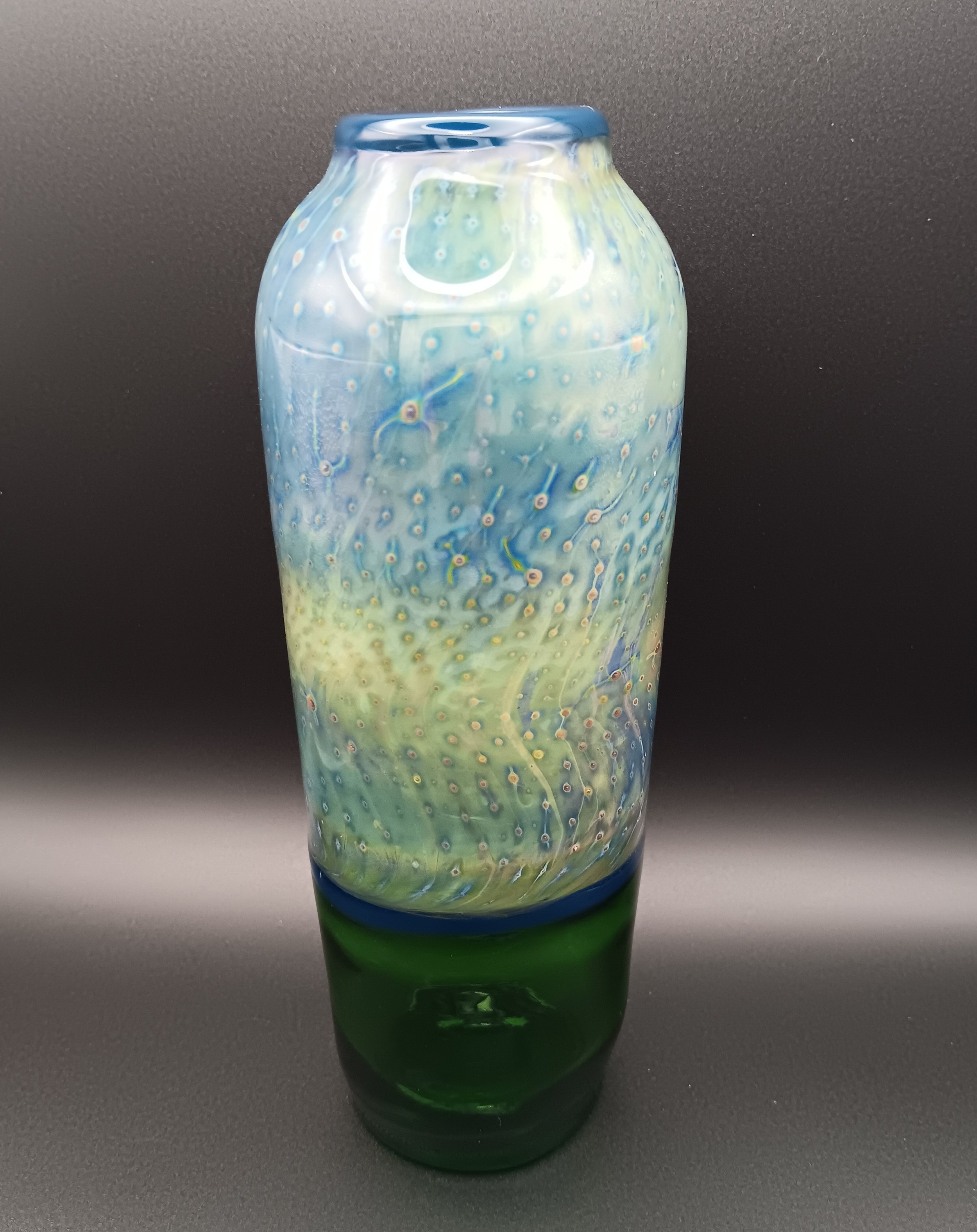 A tall glass vase with a colorful, swirling pattern of blue, green, yellow, and white, against a black background.