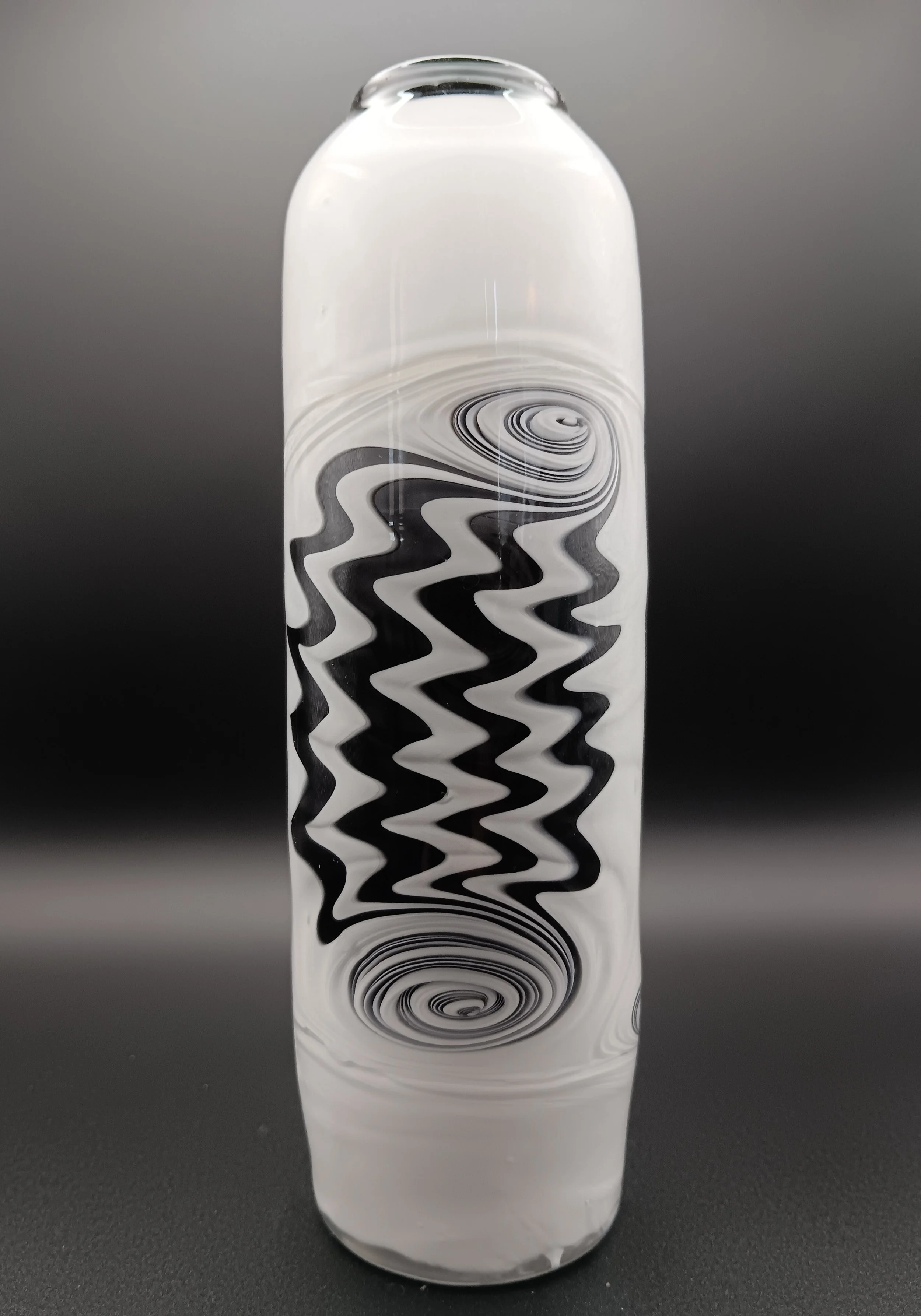 A glass vase with black and white zigzag and spiral patterns, placed on a black surface with a gray background.