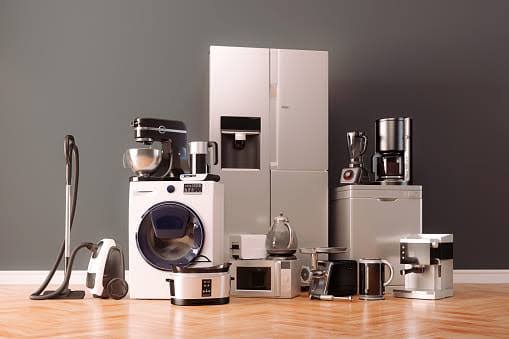 Kitchen appliances including a washing machine, refrigerator, coffee maker, and various countertop devices against a gray wall.