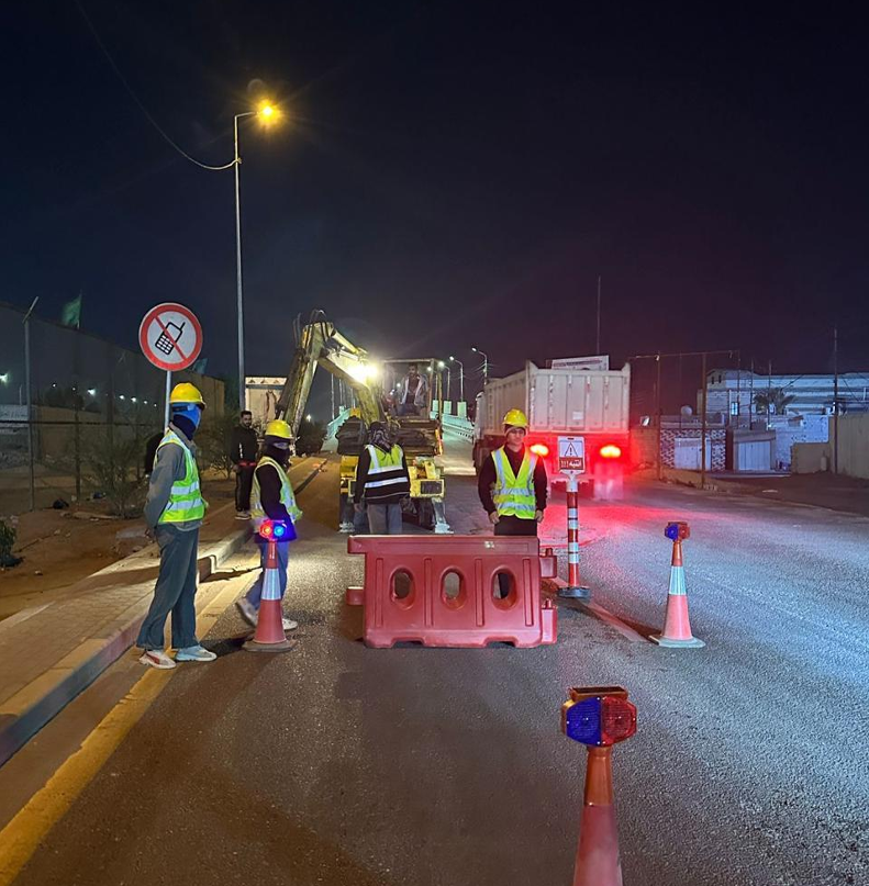 Nighttime road construction with workers in safety vests and helmets, construction equipment, traffic cones, and barriers.