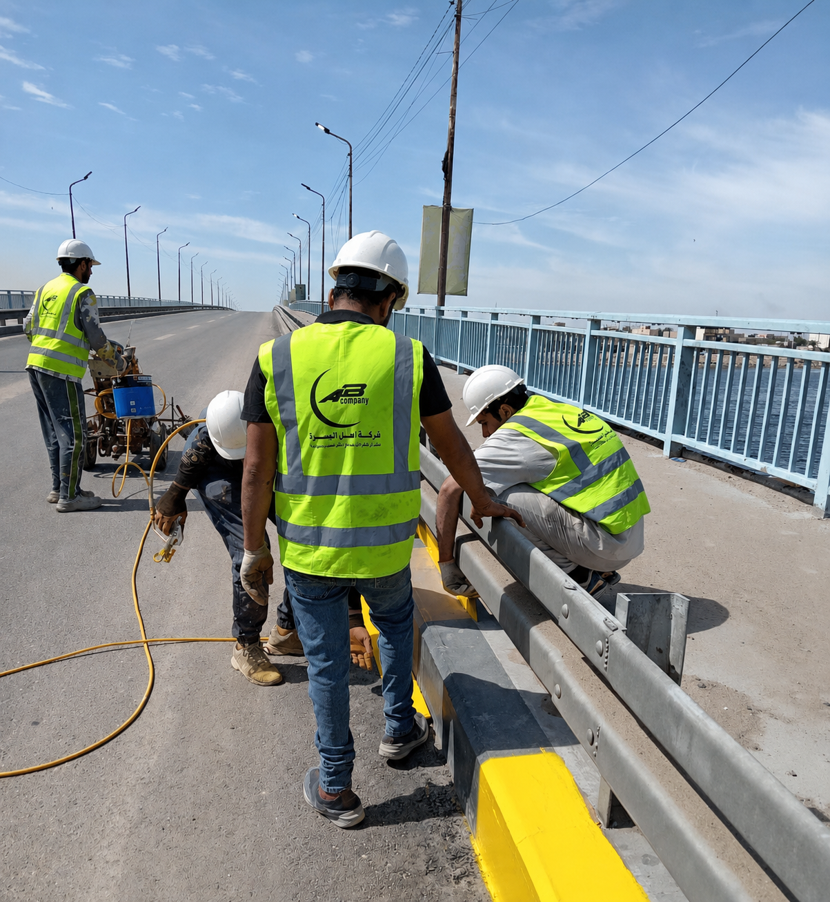 Construction workers in safety vests and helmets repair a bridge barrier on an elevated roadway over water, with utility poles and a cloudy sky in the background.