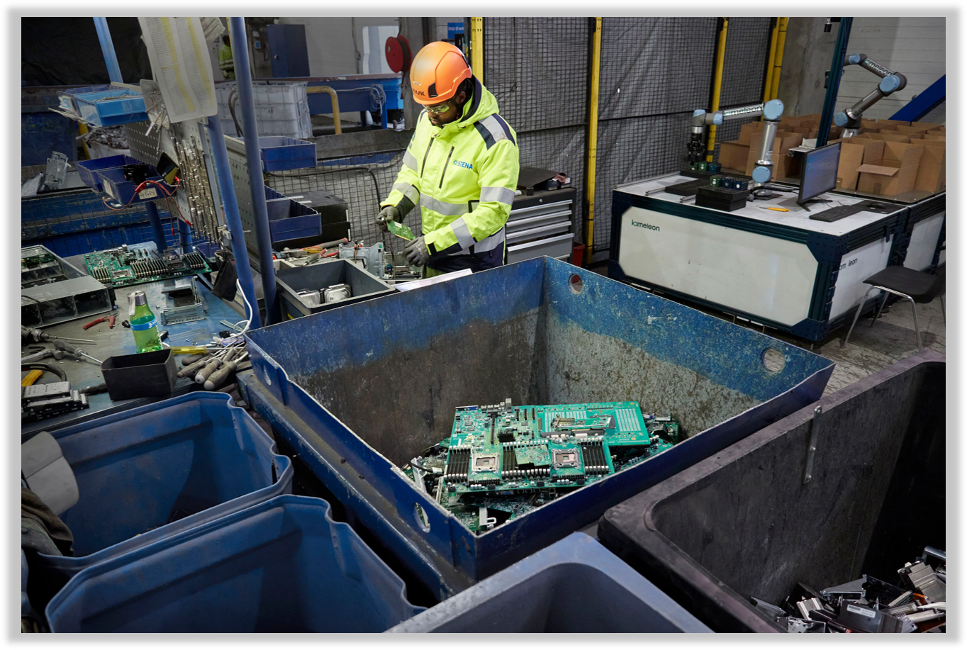 A worker in a high-visibility jacket and safety helmet sorts electronic waste in a recycling facility, with various electronic components and tools on the workbench and a large metal bin filled with circuit boards.