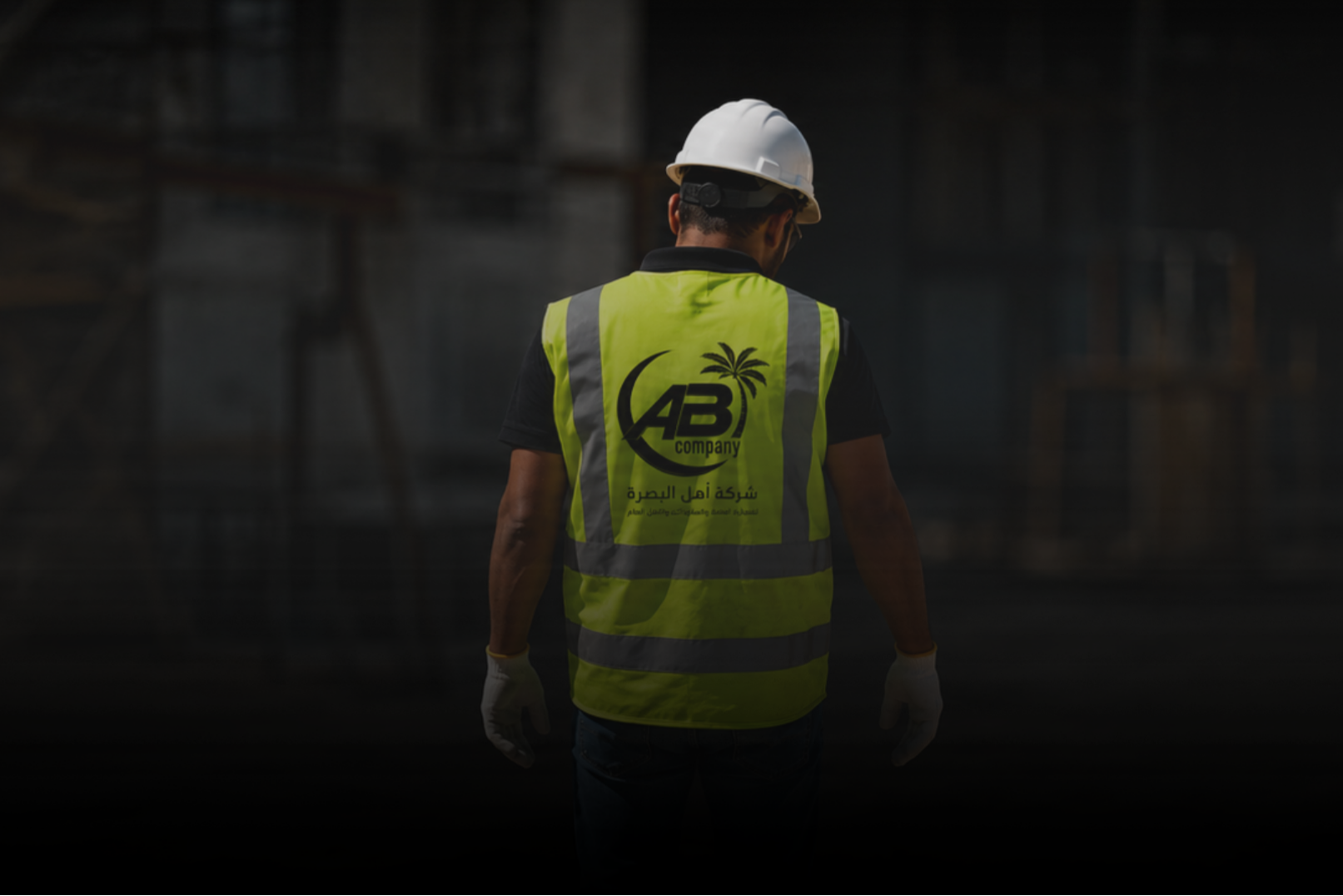 A construction worker wearing a white safety helmet and a yellow safety vest with a logo and Arabic text, standing on a construction site.