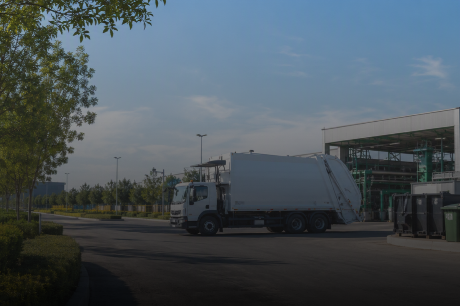 A white garbage truck parked near a recycling station outdoors on a sunny day with trees and streetlights in the background.