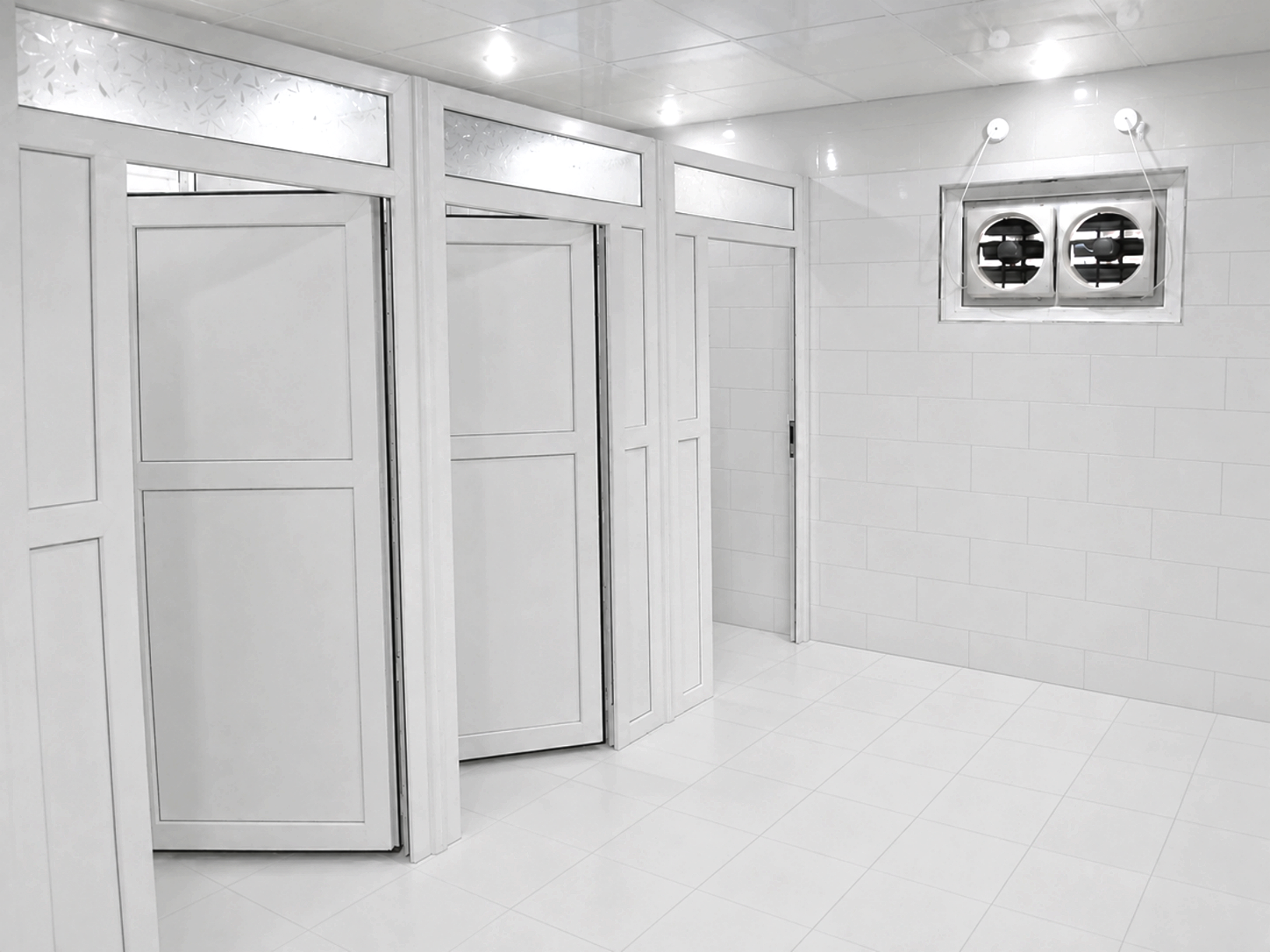 A clean, white tiled restroom with partitioned stalls and a ventilation fan high on the wall.