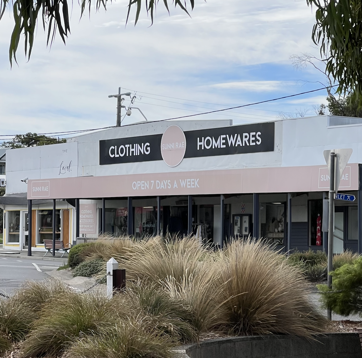 Exterior view of a clothing, homewares, and accessories store called Sunny Rae, with a sign indicating it is open 7 days a week. The store has large glass windows and door, with some items visible inside. There is a small parking lot outside, with plants and grasses in the foreground and a blue sky with clouds overhead.