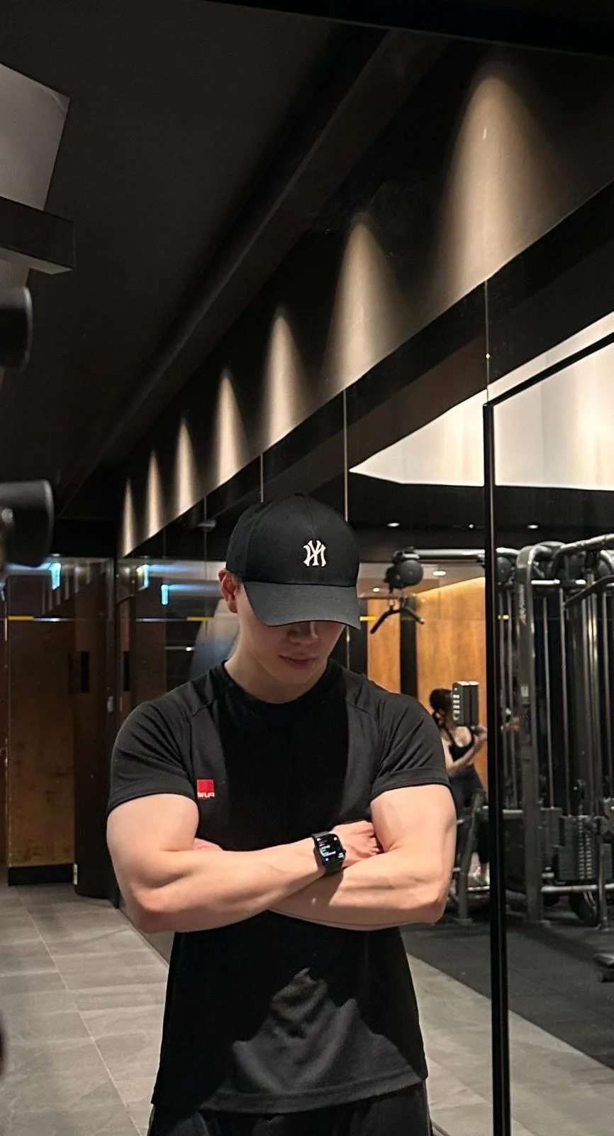 A young man with crossed arms, wearing a black baseball cap and black workout shirt, in a gym with equipment and a mirror in the background.