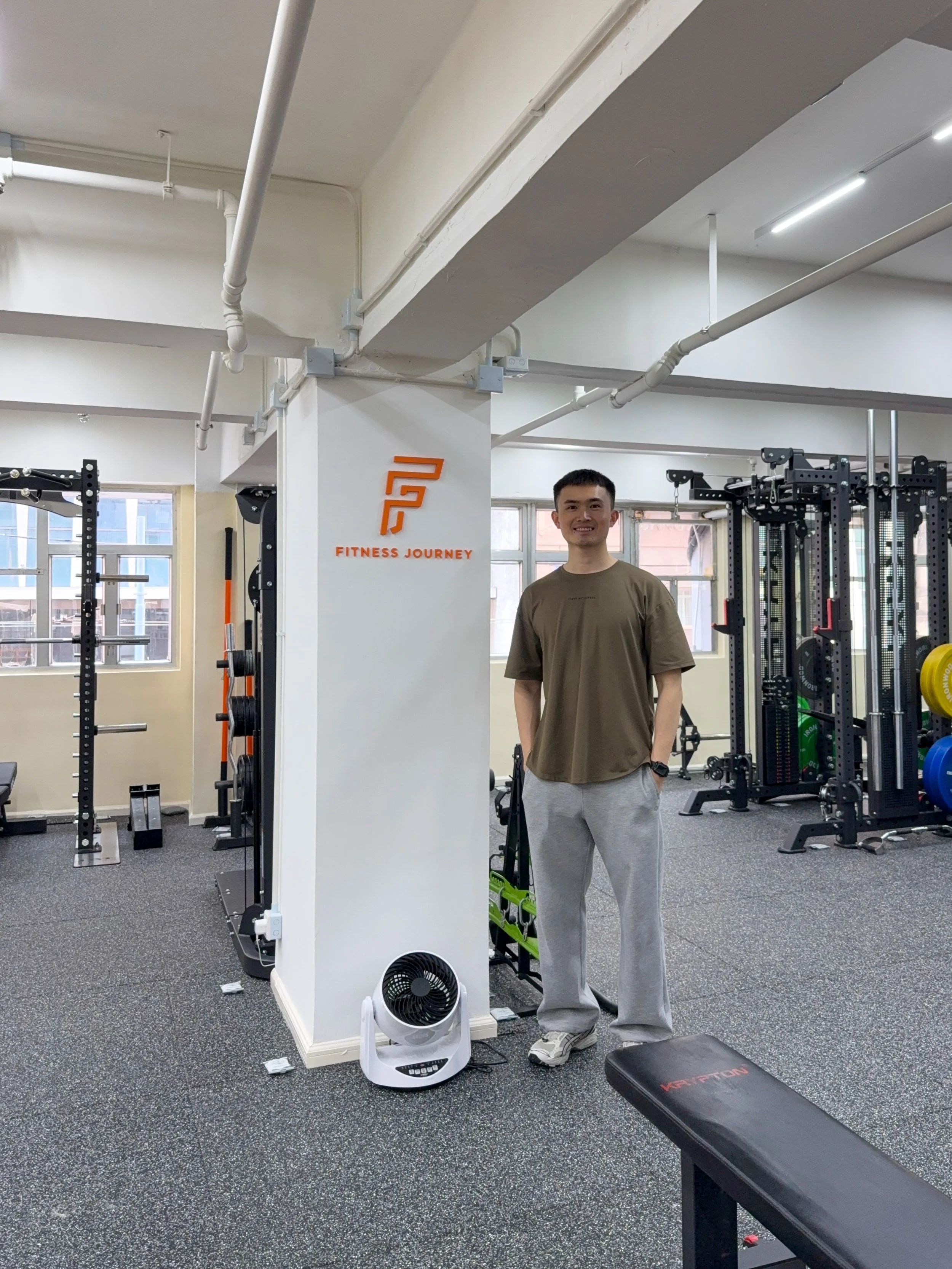 A young man standing in a gym, wearing a brown T-shirt and gray sweatpants, smiling, with gym equipment including a squat rack, weights, and a ventilation fan nearby. The gym has large windows and a sign that reads 'Fitness Journey.'
