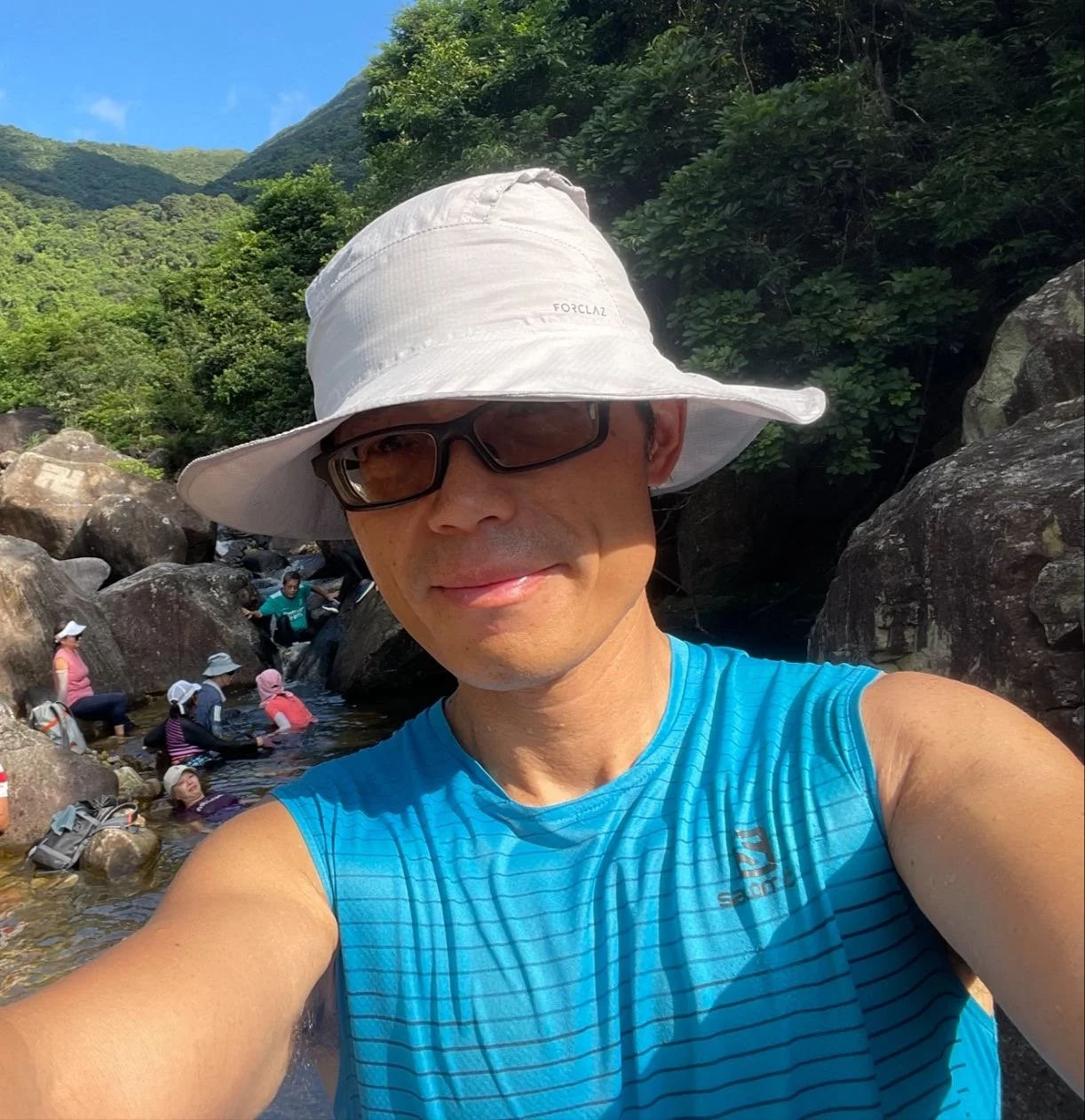 A man wearing a white sun hat, sunglasses, and a blue sleeveless shirt taking a selfie by a rocky stream in a lush green forest with a mountain in the background, while several people wade in the water.