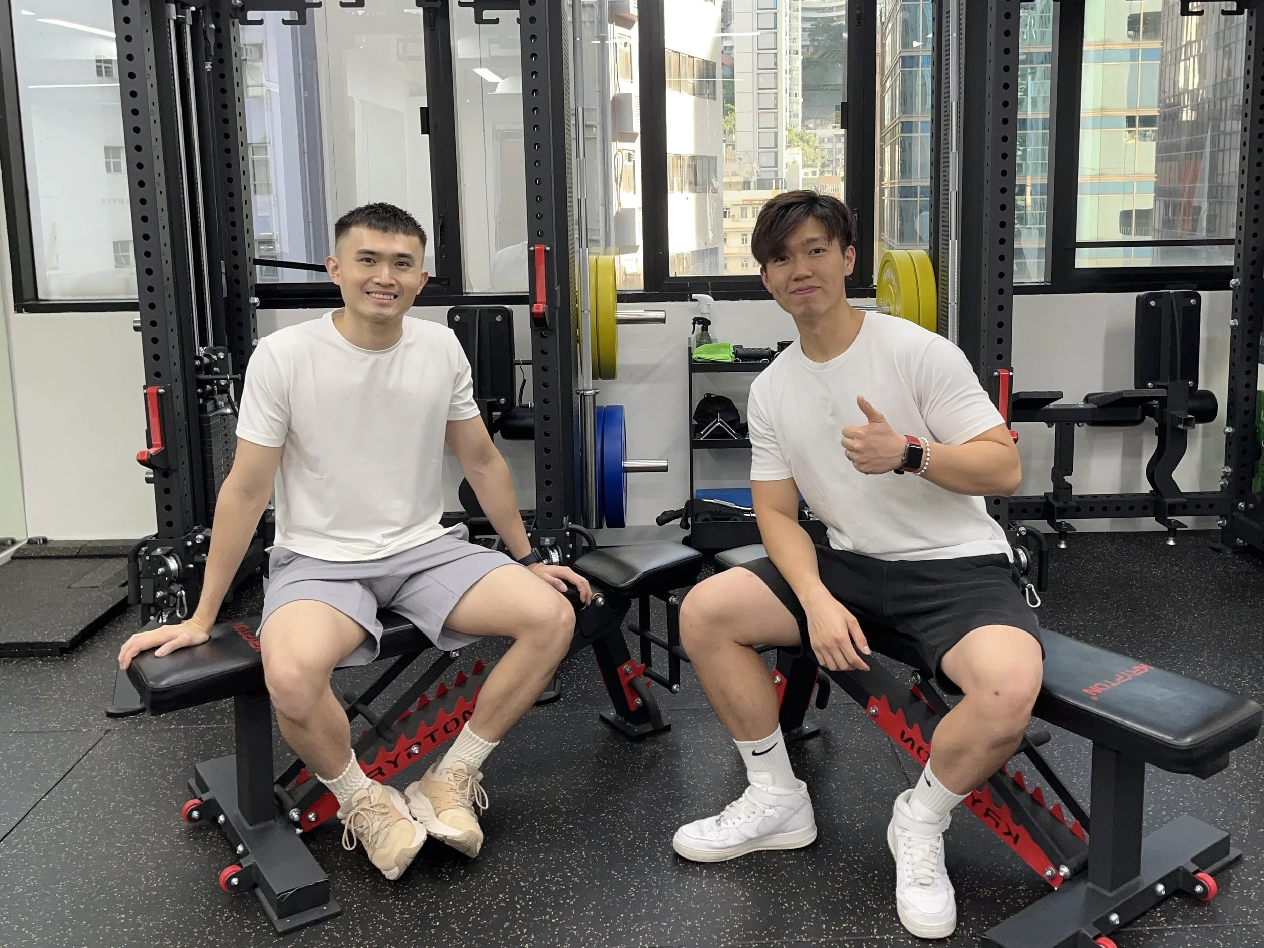 Two young men sitting inside a gym near weightlifting equipment, smiling and making a thumbs-up gesture.