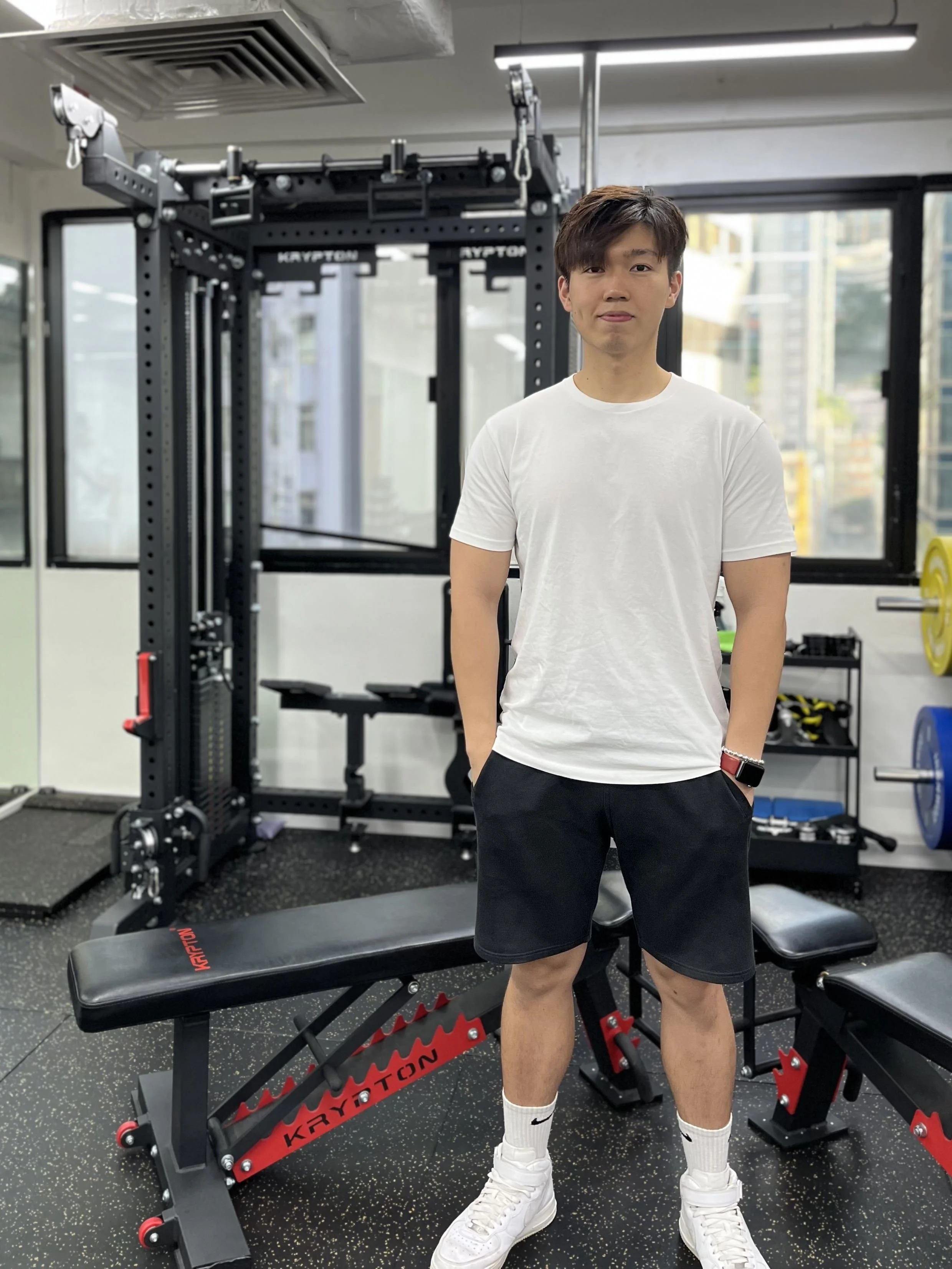 A young man standing in a gym, wearing a white t-shirt, black shorts, and white sneakers, with gym equipment in the background including a black weightlifting bench and a power rack.