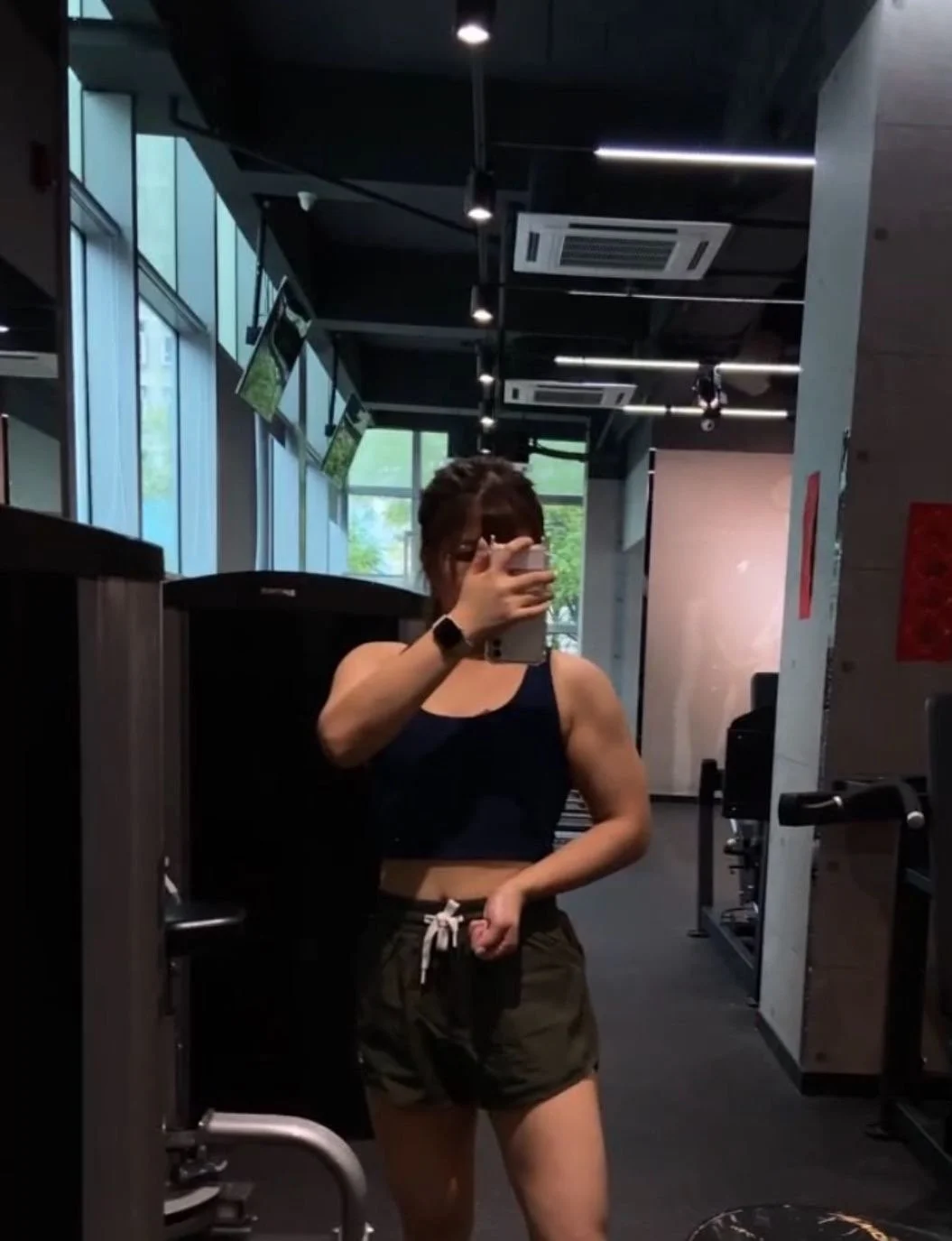 A woman in workout clothes taking a mirror selfie in a gym with large windows, fitness equipment, and TV screens visible in the background.