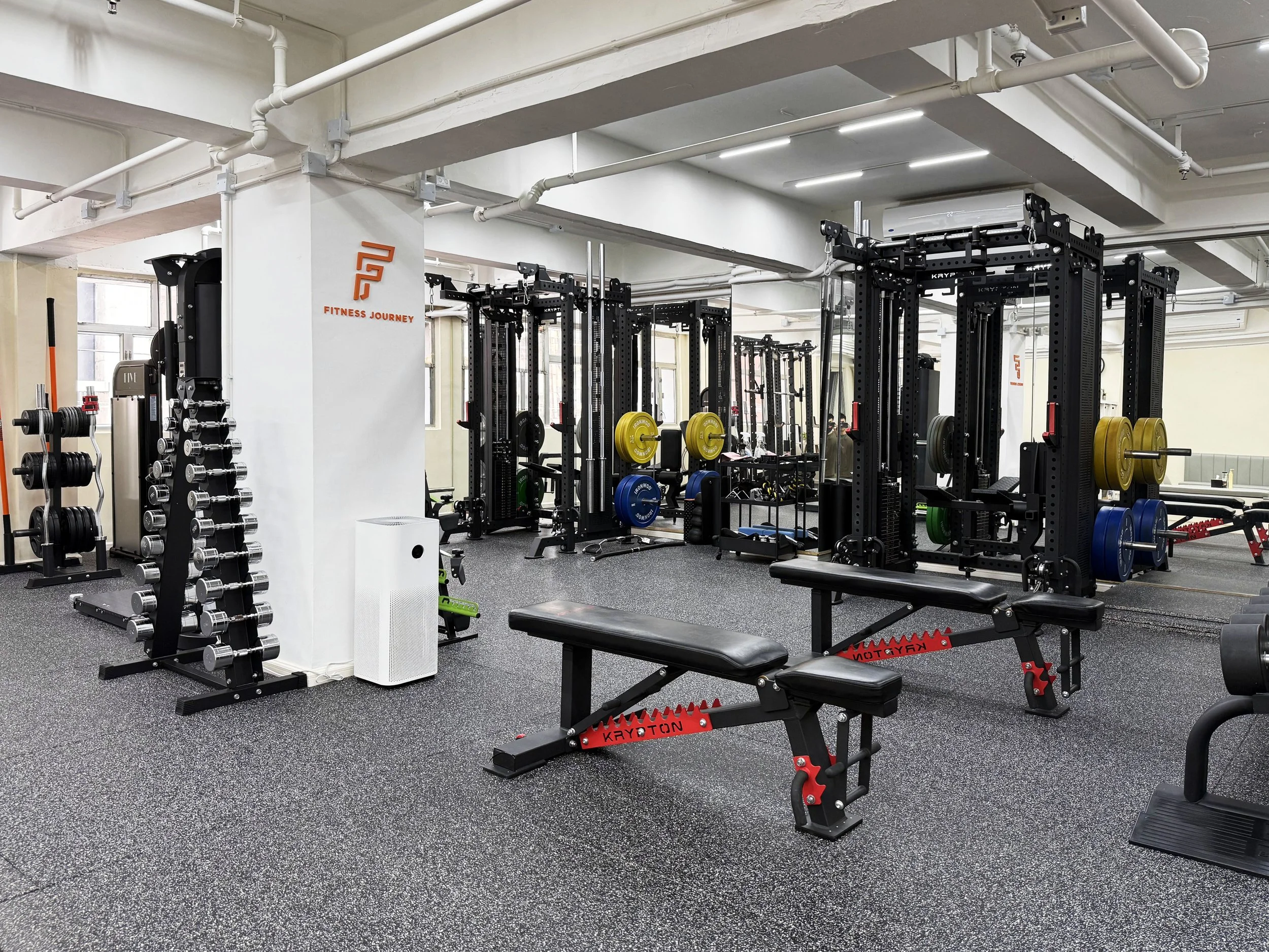 Fitness gym with weightlifting equipment, including free weights, weight machines, and benches, in a bright room with large windows and industrial ceiling pipes.
