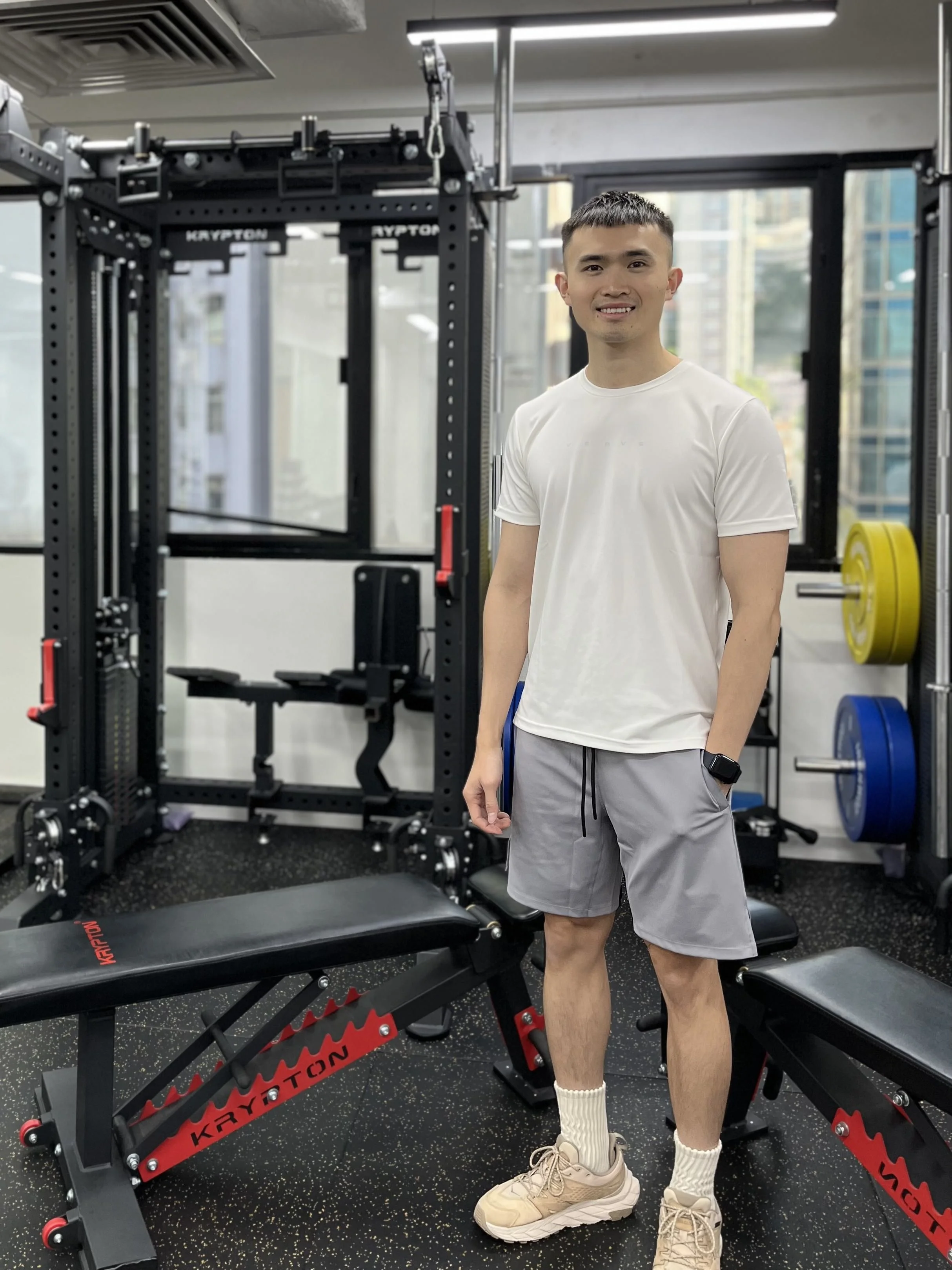 A young man in athletic clothes and sneakers smiling at the camera inside a gym near workout equipment, including a weight rack, a Bench, and weight plates.