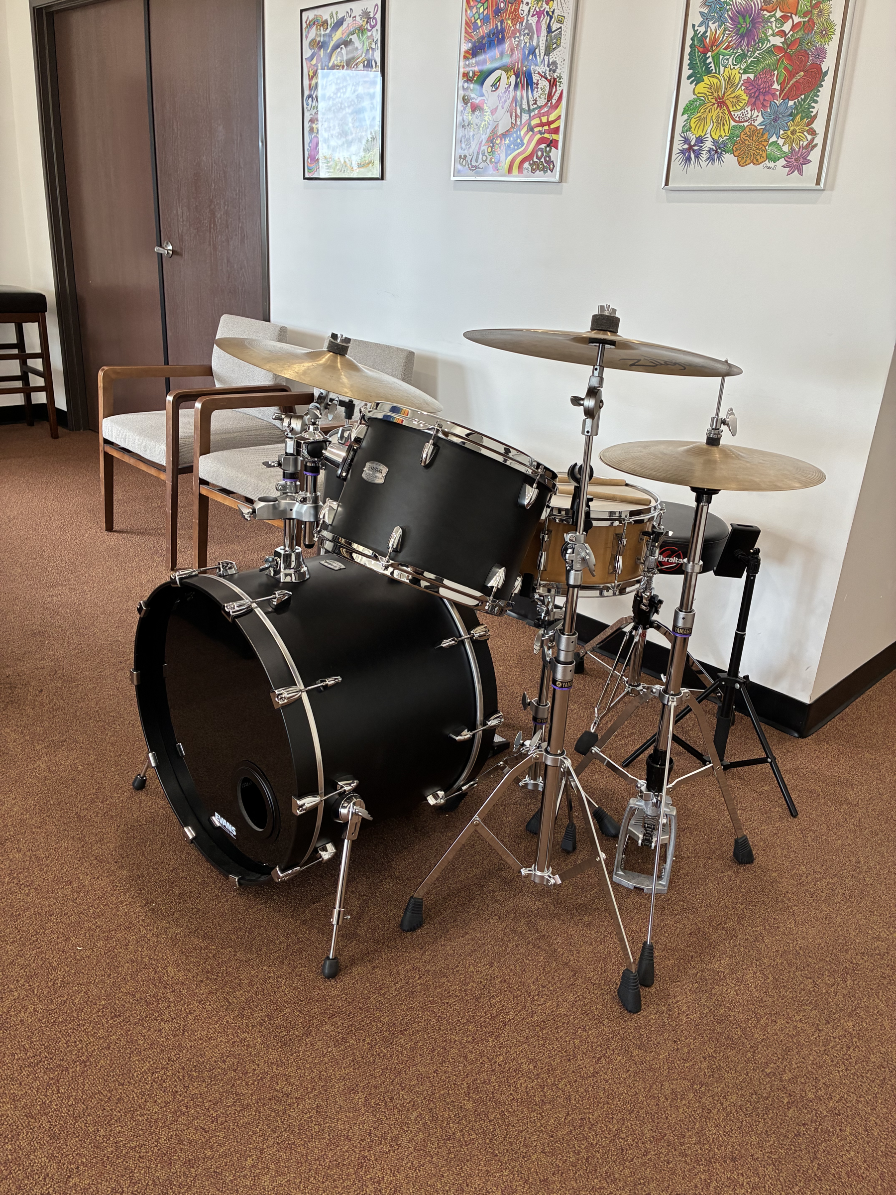 A black drum set with cymbals on stands is positioned on a beige carpeted floor in a room, with two wooden chairs and a white wall decorated with three colorful framed art pieces, one featuring an abstract design and the other a floral pattern.