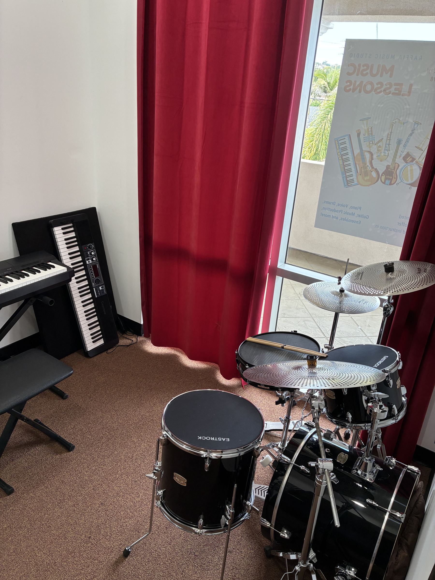 A room with musical instruments including a black electronic keyboard, a digital drum set, and a poster on the window advertising a music lesson class.