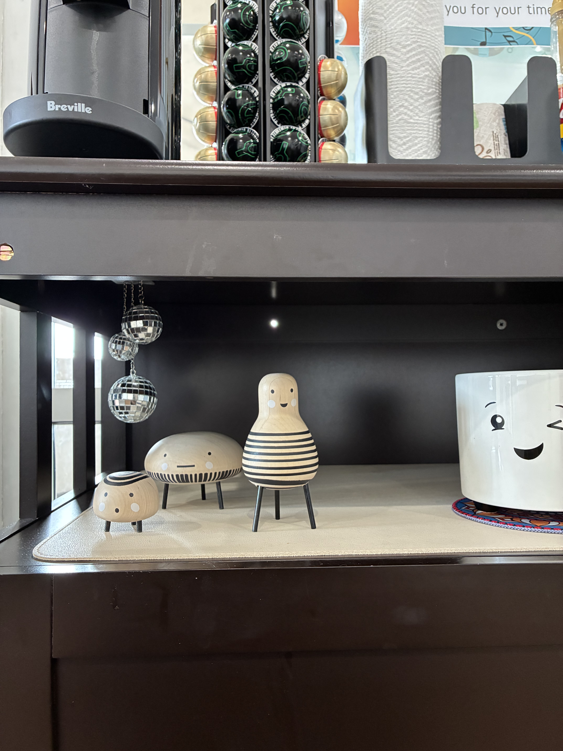 Display shelf with decorative toys including a group of wood-shaped figures with smiley faces and black and white stripes, hanging disco balls, and various kitchen items.