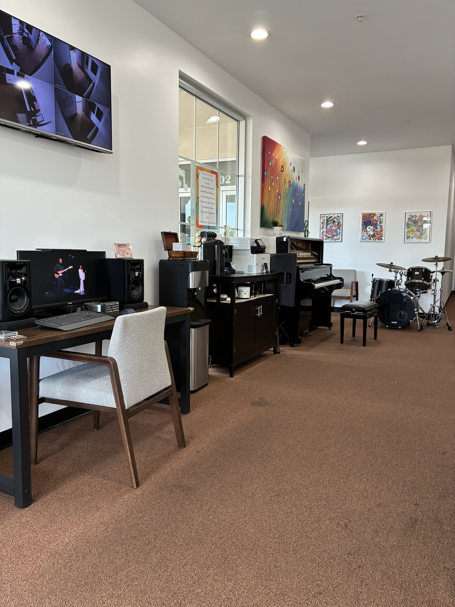 An interior room with a computer desk, piano, drums, and colorful artwork on the wall.