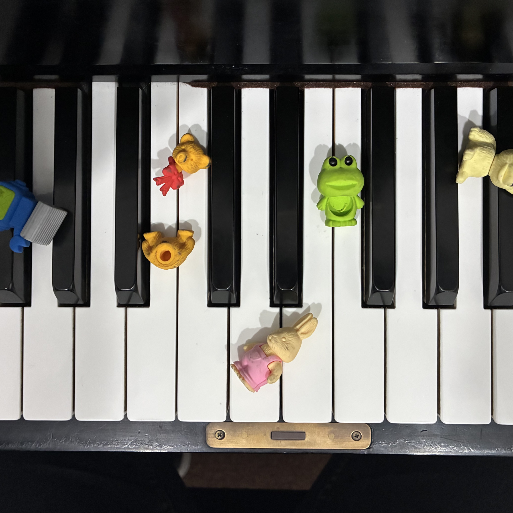 A piano keyboard with small plastic animal toys placed on the keys, including a yellow monkey, a green frog, and a cream-colored rabbit.