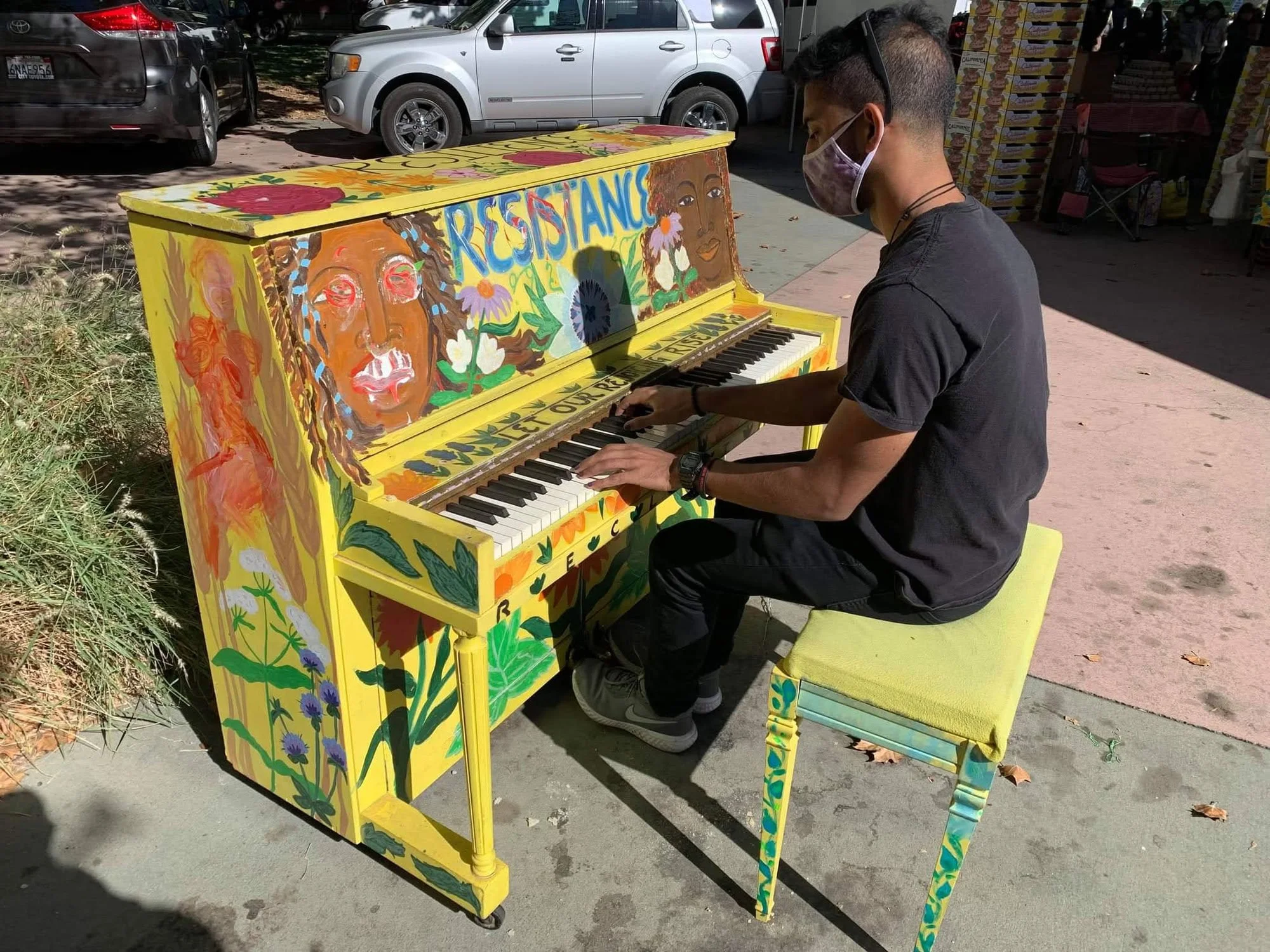 A man playing a colorful, hand-painted piano on a sidewalk. The piano has vibrant floral and portrait artwork, with the word 'RESISTANCE' painted on it.