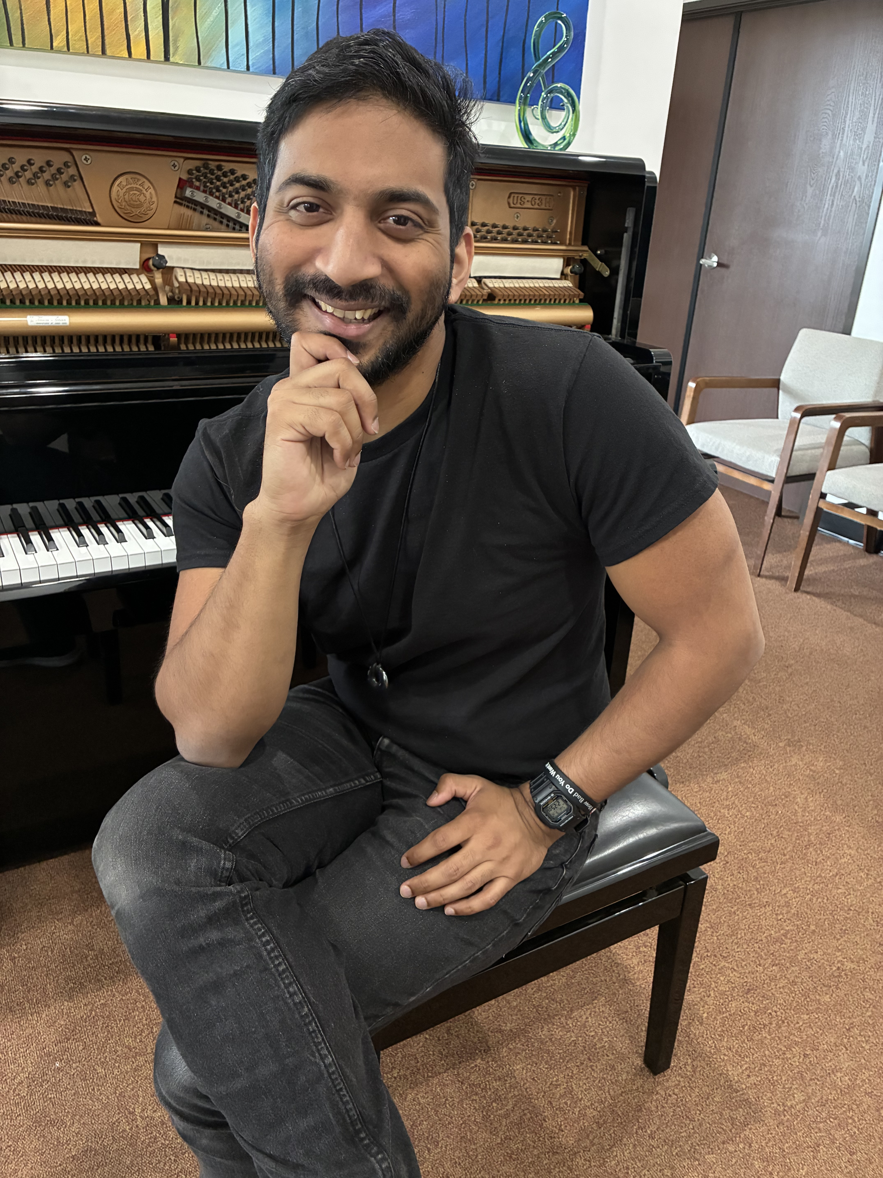 A smiling man with dark hair and a beard sitting in front of an open upright piano, wearing a black t-shirt, a wristwatch, and a bracelet, with colorful wall art and chairs in the background.