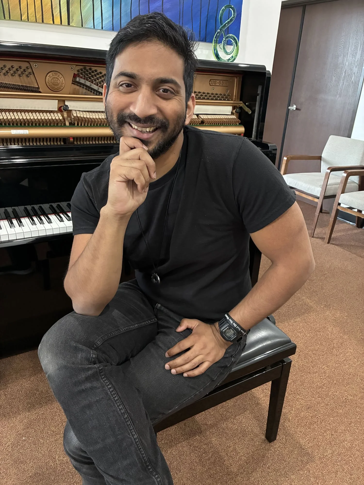 A smiling man sitting on a piano bench in front of an open piano, with a colorful painting and chairs in the background.