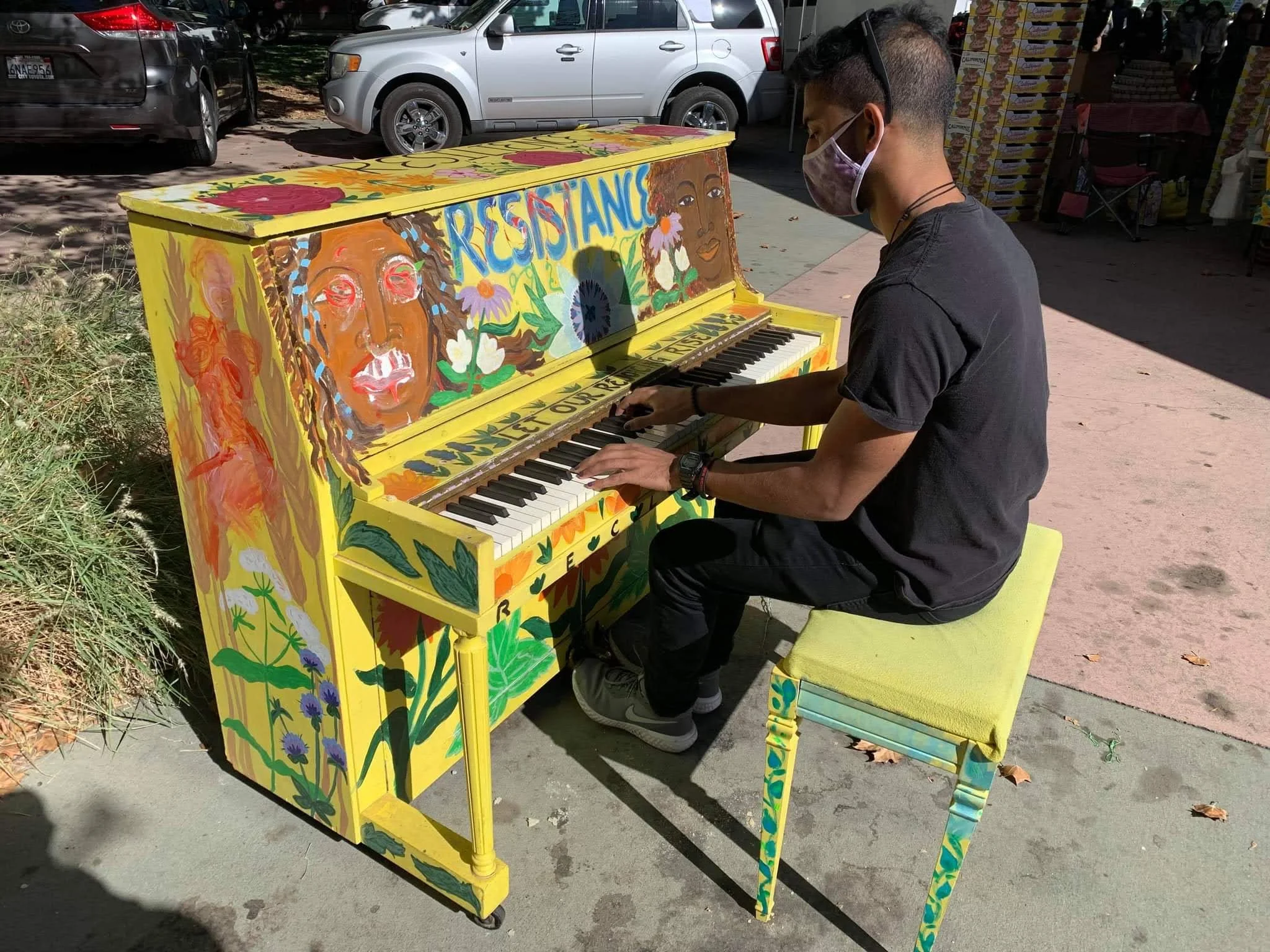 A person wearing a face mask playing a brightly painted yellow piano with floral and face designs outdoors, with parked cars and a market stall in the background.