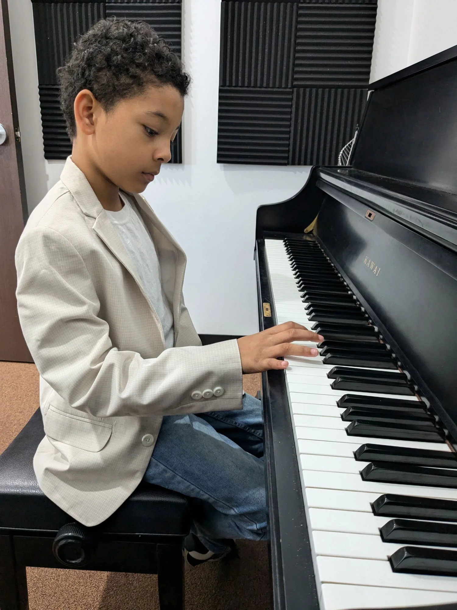 A young boy wearing a beige blazer and jeans sitting at a black Kawai piano, playing it with focus 