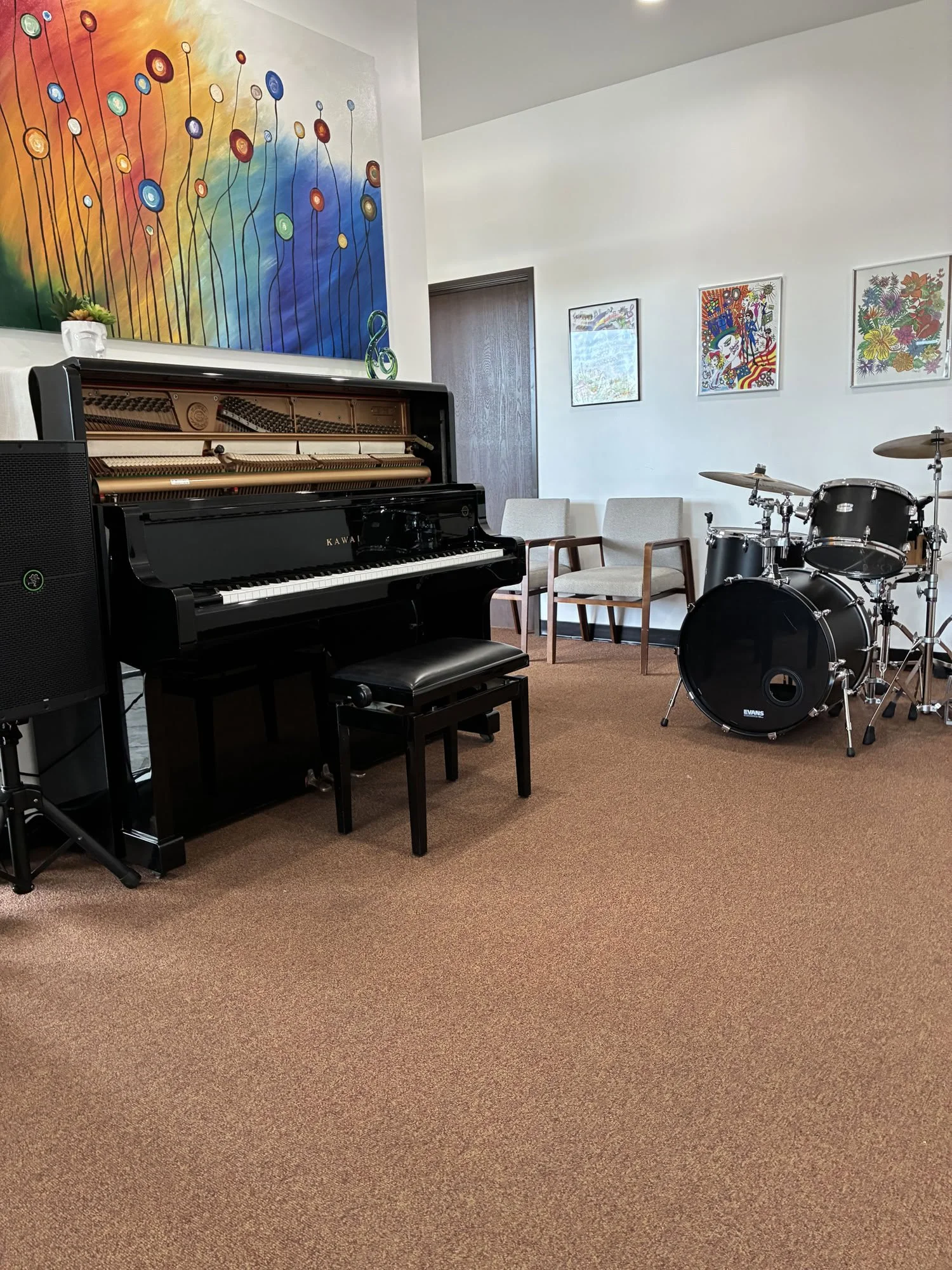 A room with a colorful abstract painting on the wall, a black upright piano with a bench, chairs, and a drum set.