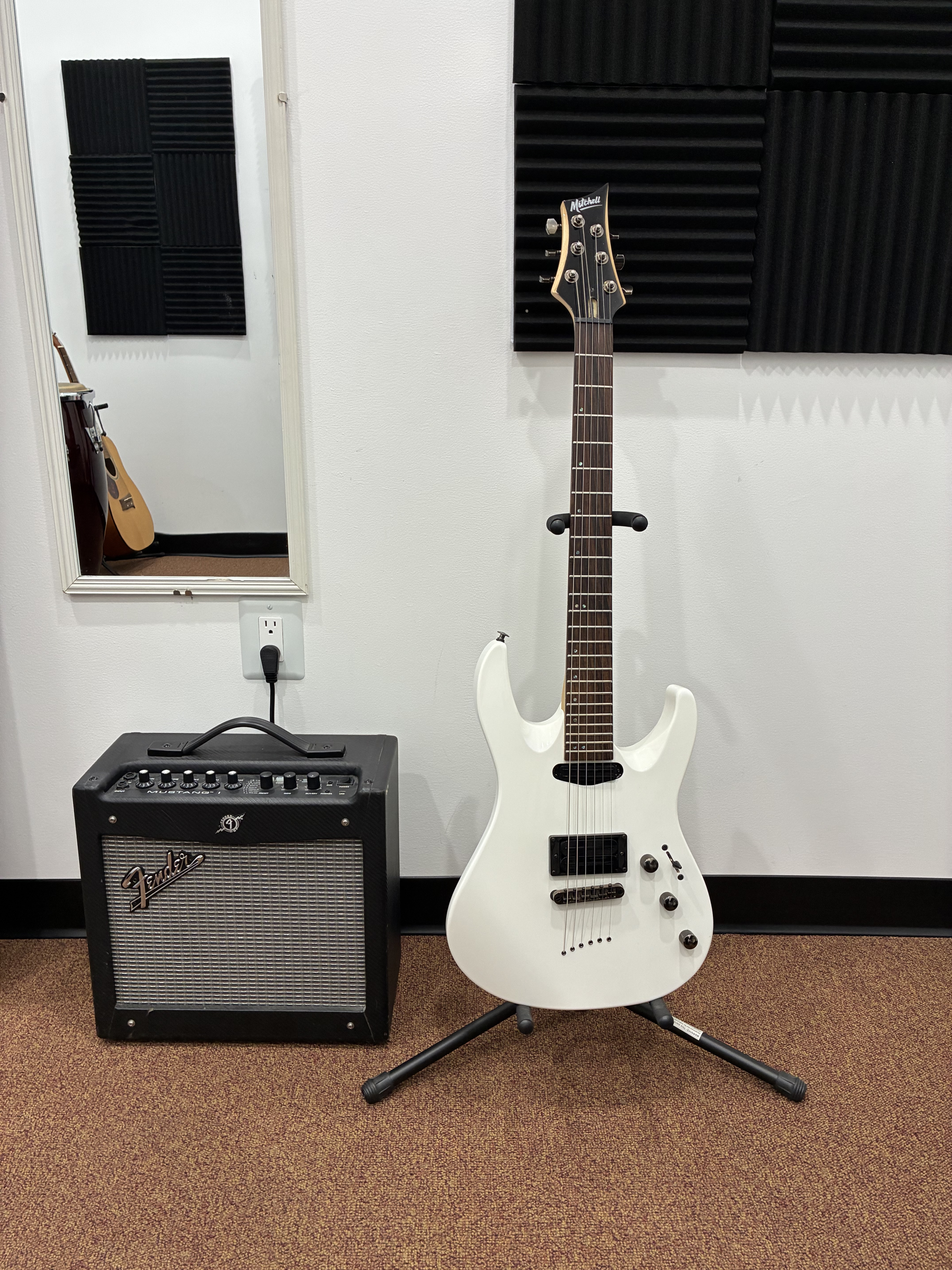 White electric guitar on stand, positioned in front of black soundproofing foam on a wall, with a Fender amplifier on a brown carpeted floor.