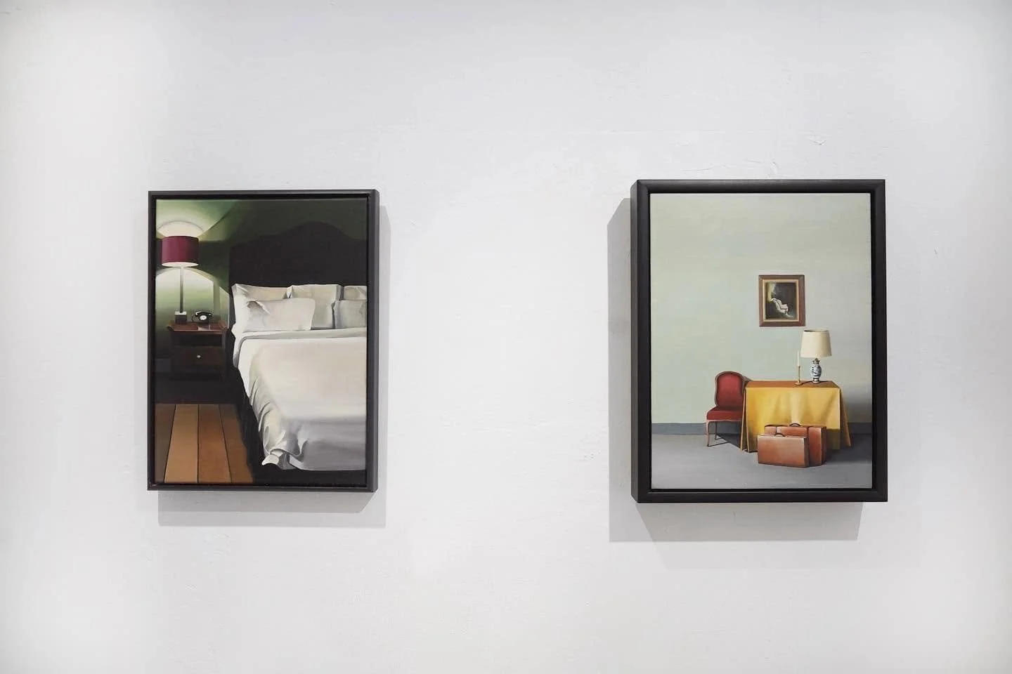 Two framed paintings hanging on a white gallery wall. The left painting depicts a bedroom with a neatly made bed, a nightstand with a lamp and a telephone, and wooden floorboards. The right painting depicts a minimalist room with a small table draped
