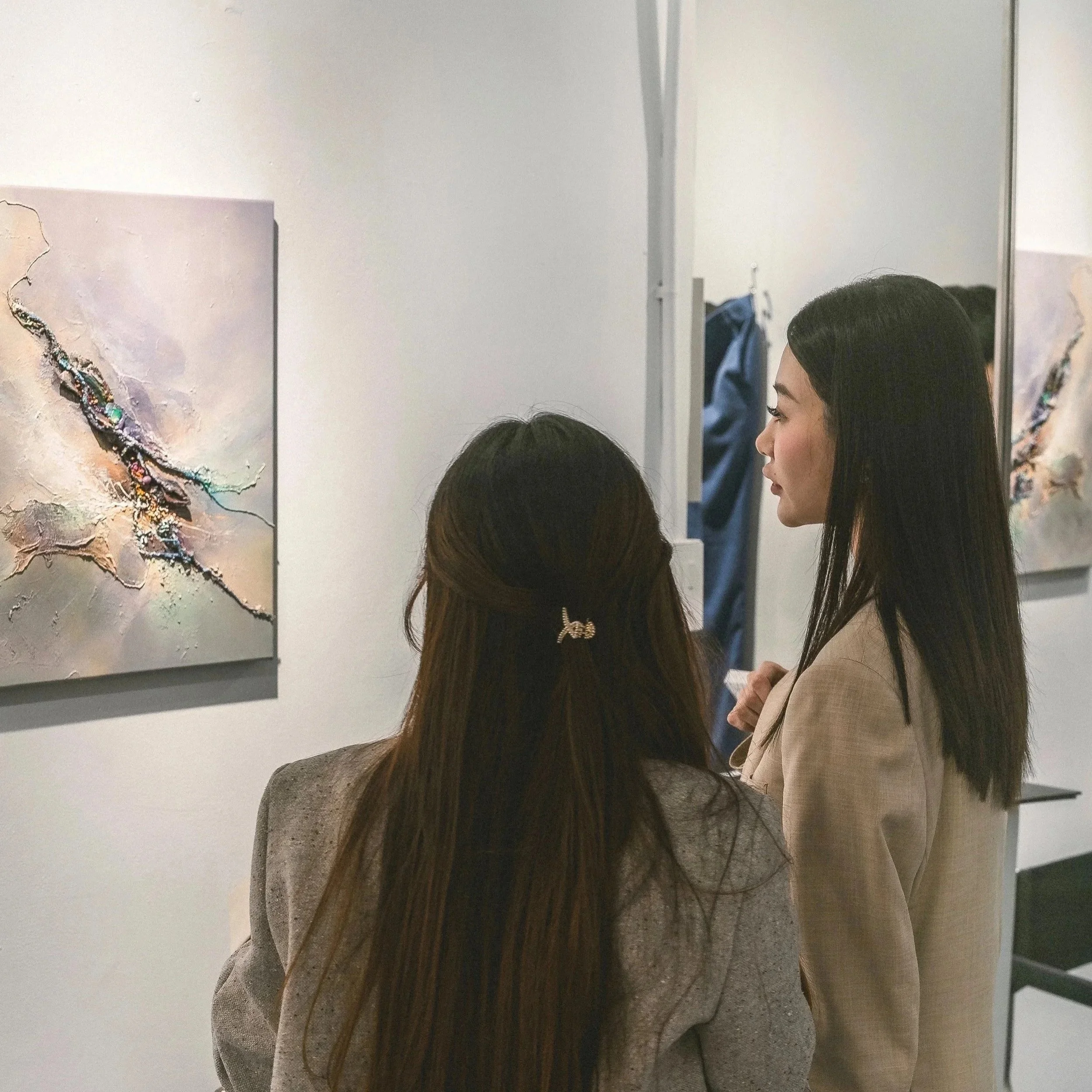 A group of people, including a woman with long brown hair and another with dark hair and a hair clip, looking at an abstract painting in an art gallery.